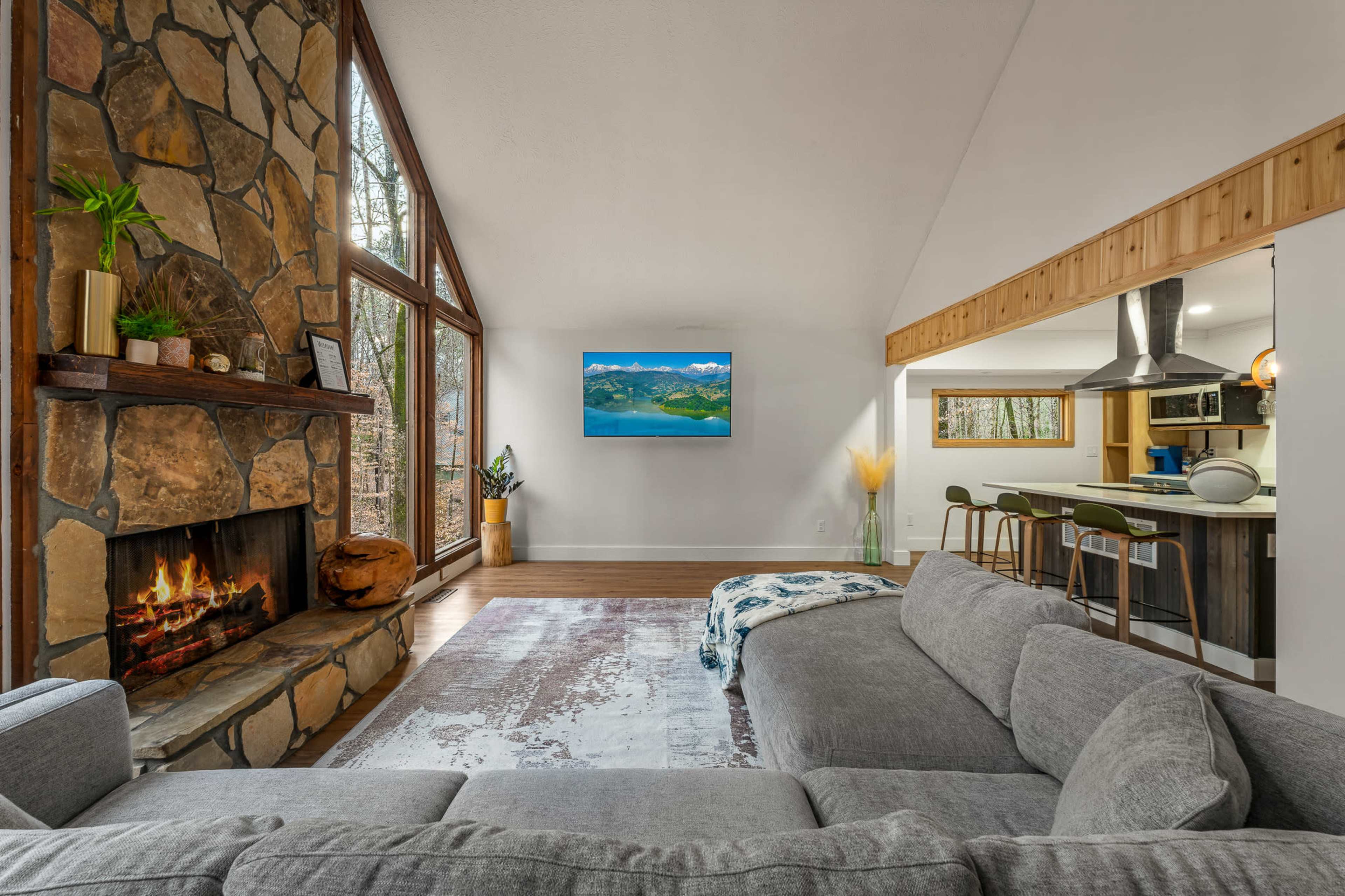 A modern living room features a stone fireplace, large windows overlooking a wooded area, and a wall-mounted television.