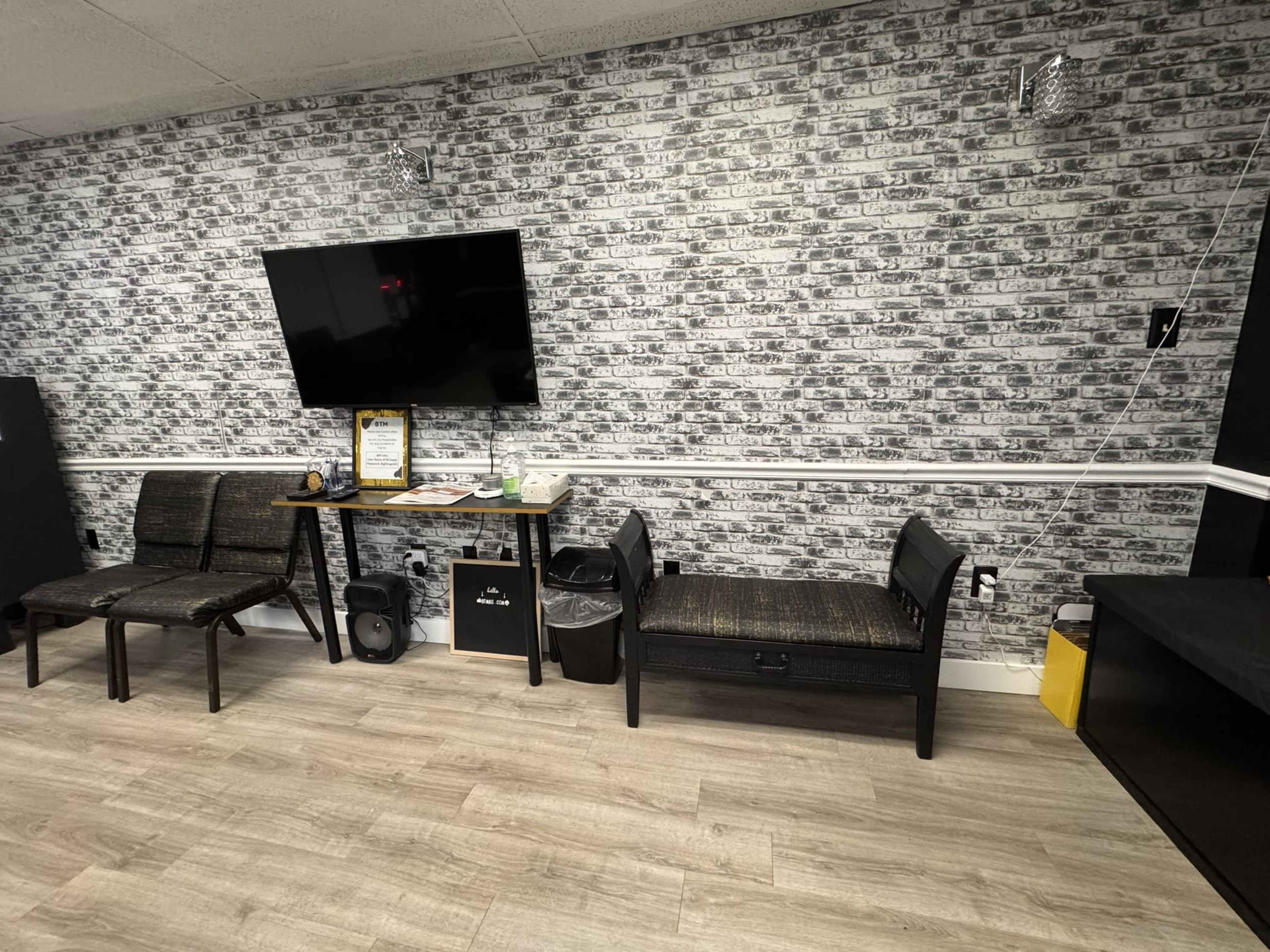 The image shows a room with a gray brick-patterned wall, a wall-mounted television, a table with some items on it, and two black chairs.