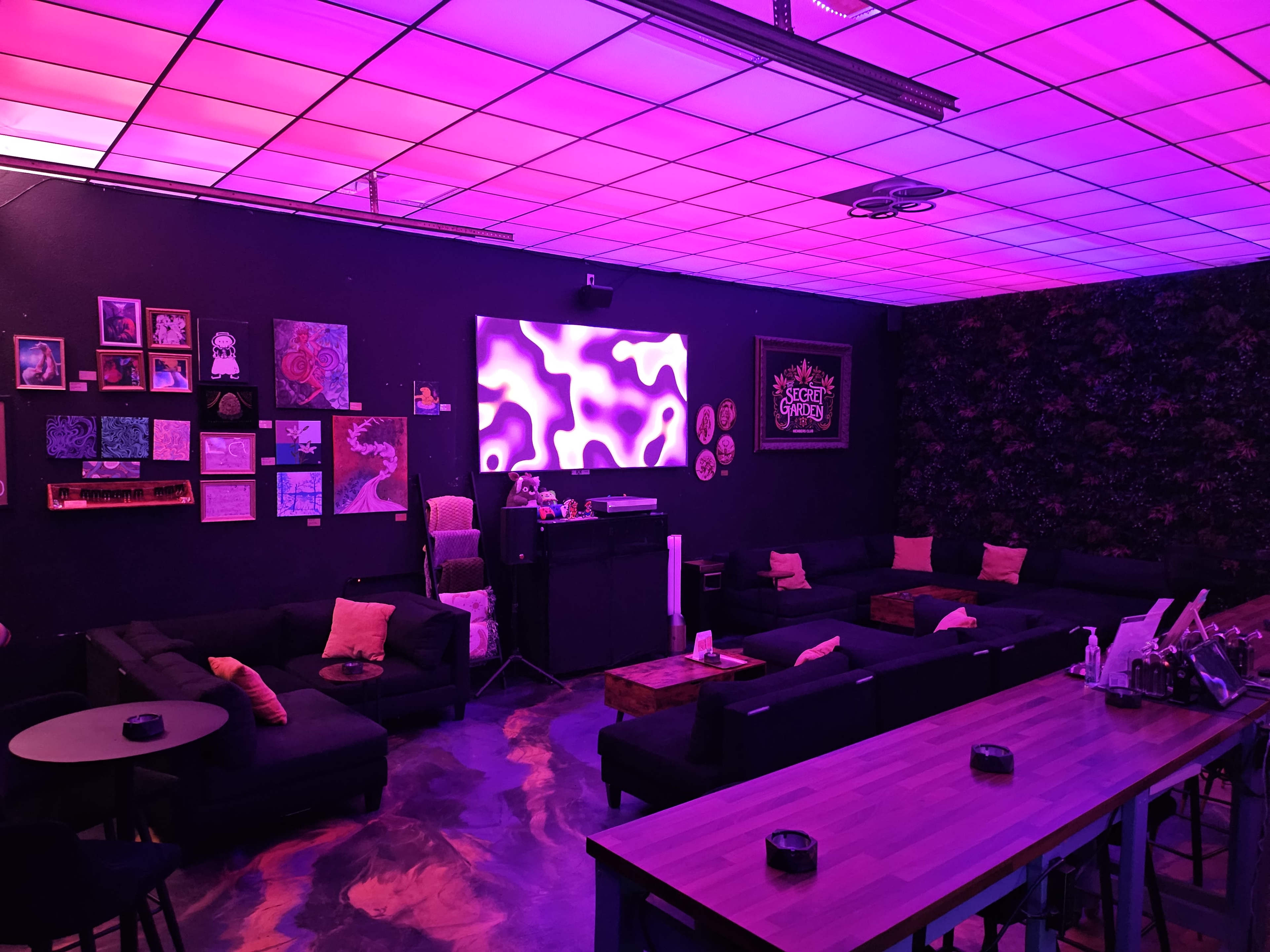 The image shows a modern lounge area with dark sofas, a large screen displaying colorful graphics, and dim purple lighting, accentuated by various artworks on the walls.