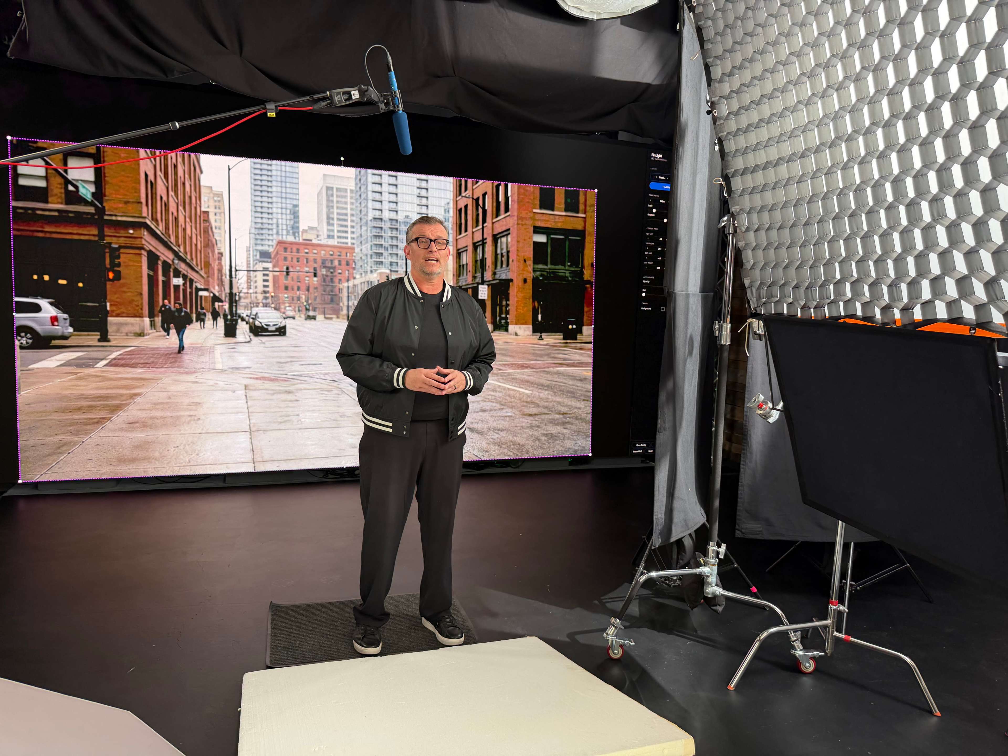 A man stands in a studio setting with a large digital screen displaying an urban street scene behind him.