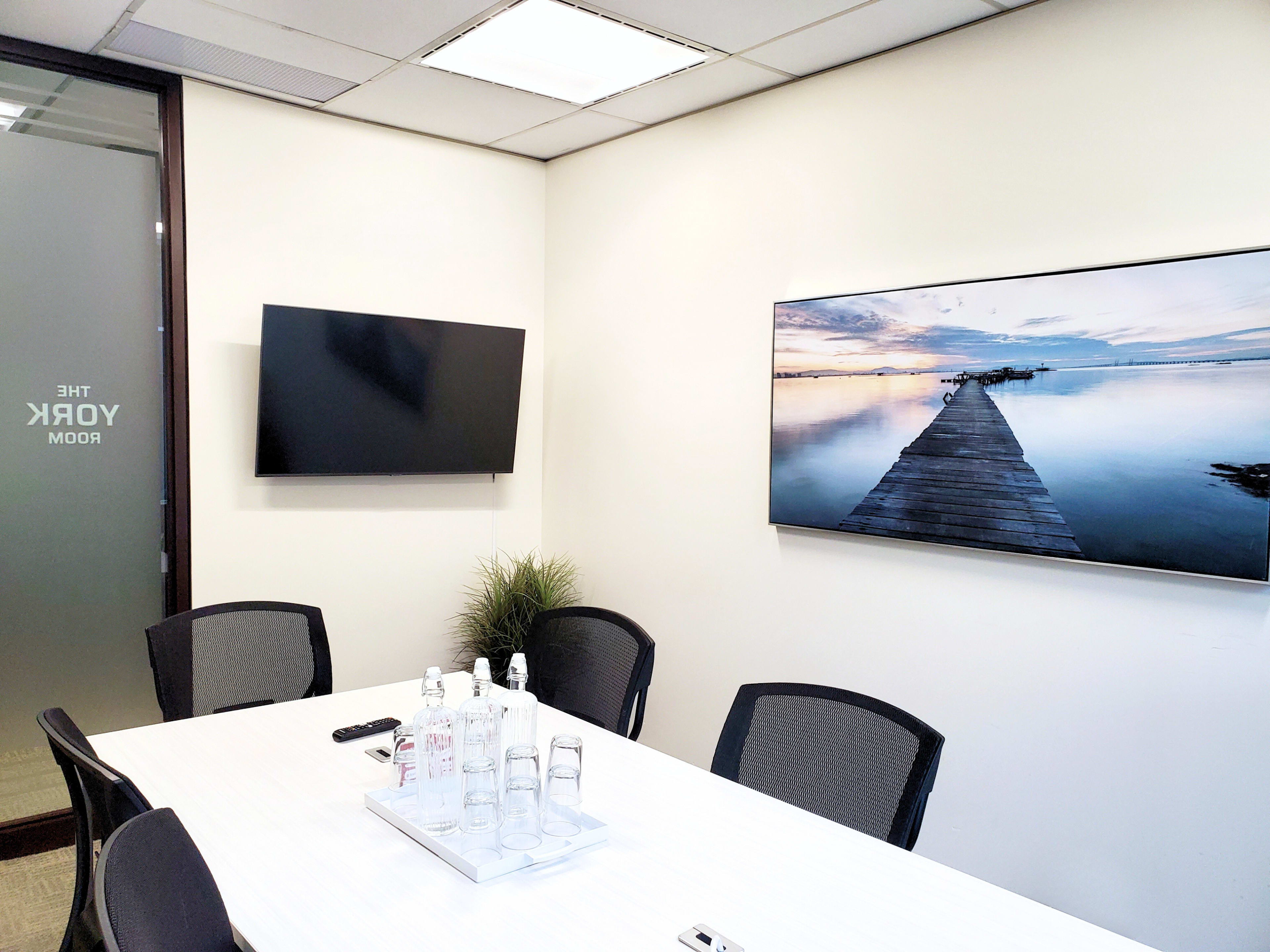 The image shows a conference room with a long table, several black chairs, a large television mounted on the wall, and a scenic photo of a pier over water.