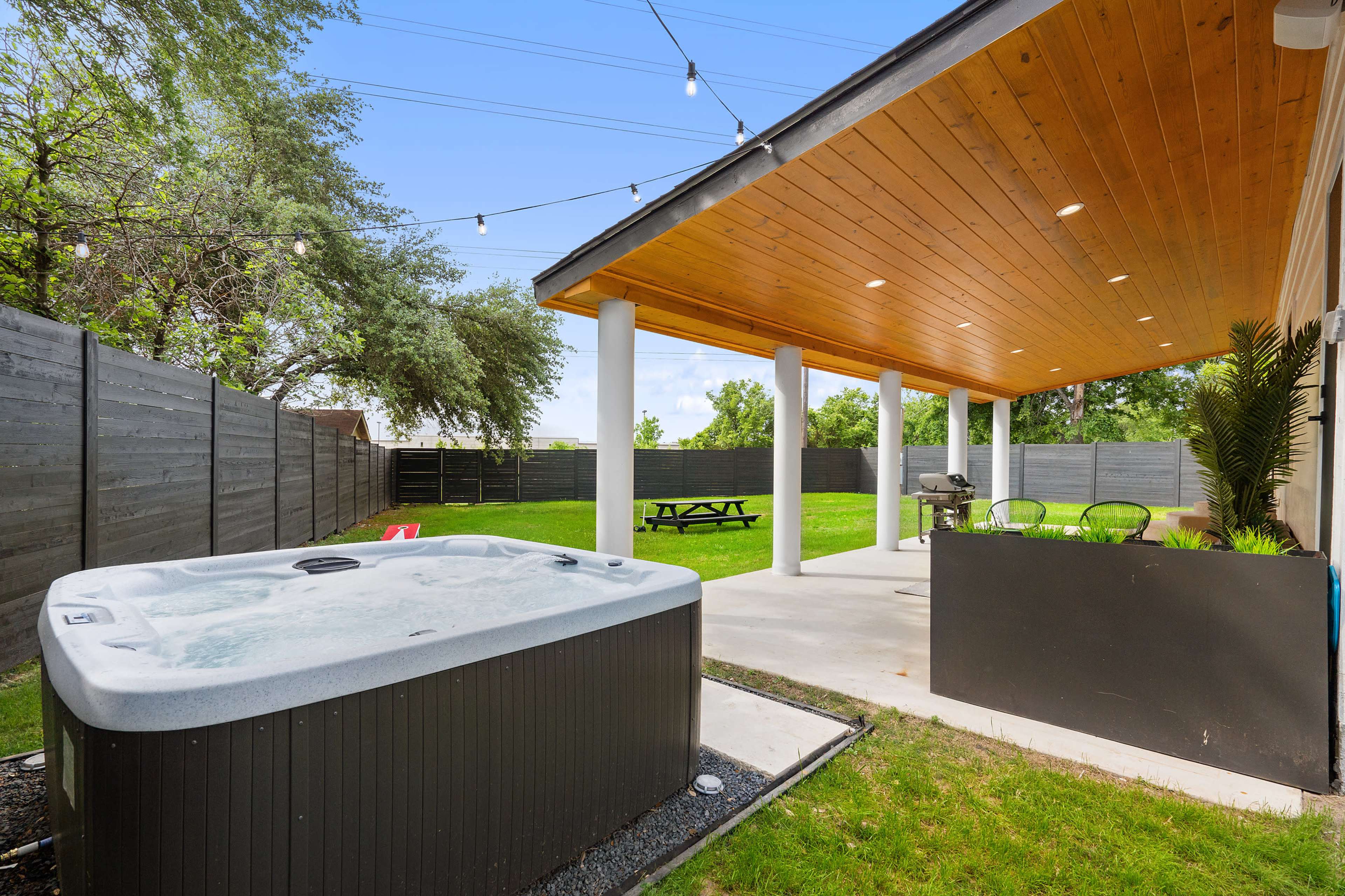 A backyard patio with a hot tub, a picnic table, and a grill area under a wooden roof, surrounded by a green lawn and a wooden fence.