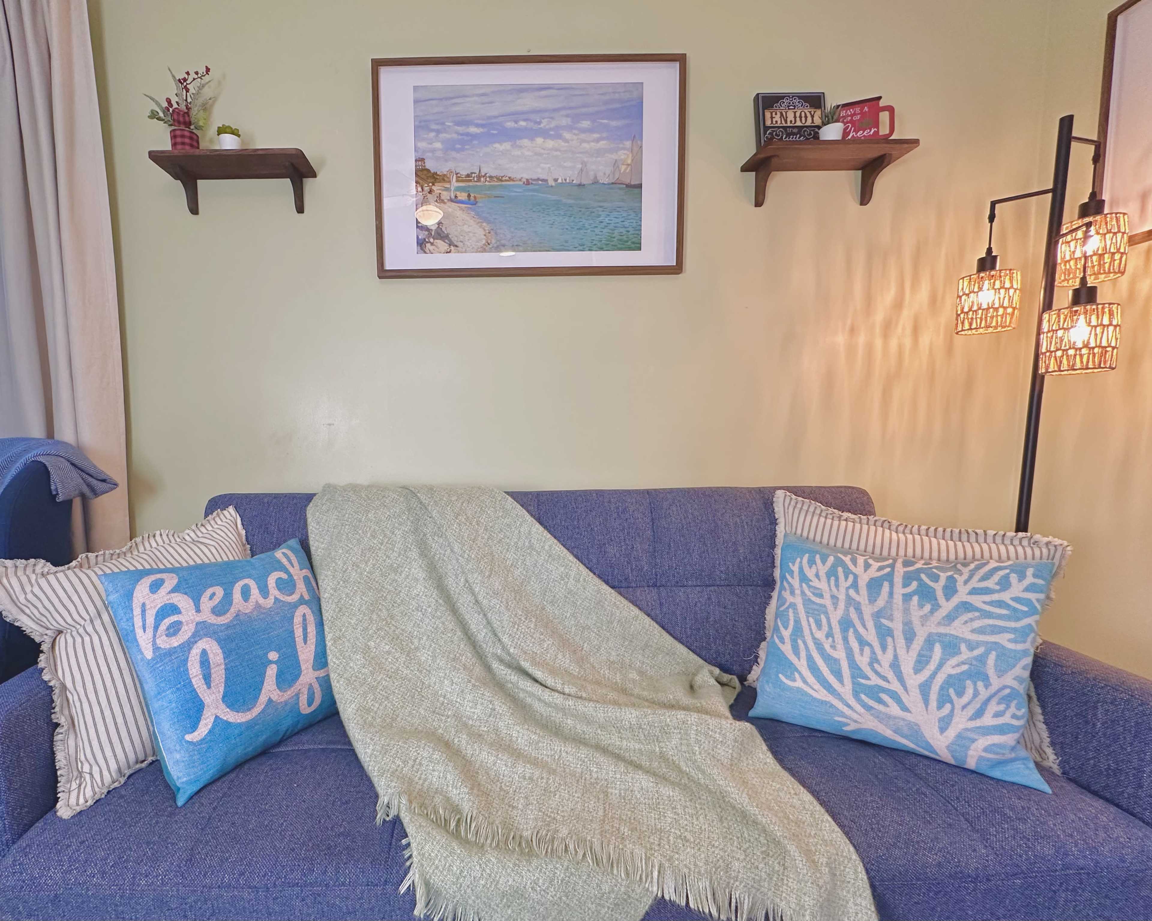 A blue sofa with decorative pillows and a throw blanket is positioned against a light green wall, featuring a framed picture and shelves with decorative items.