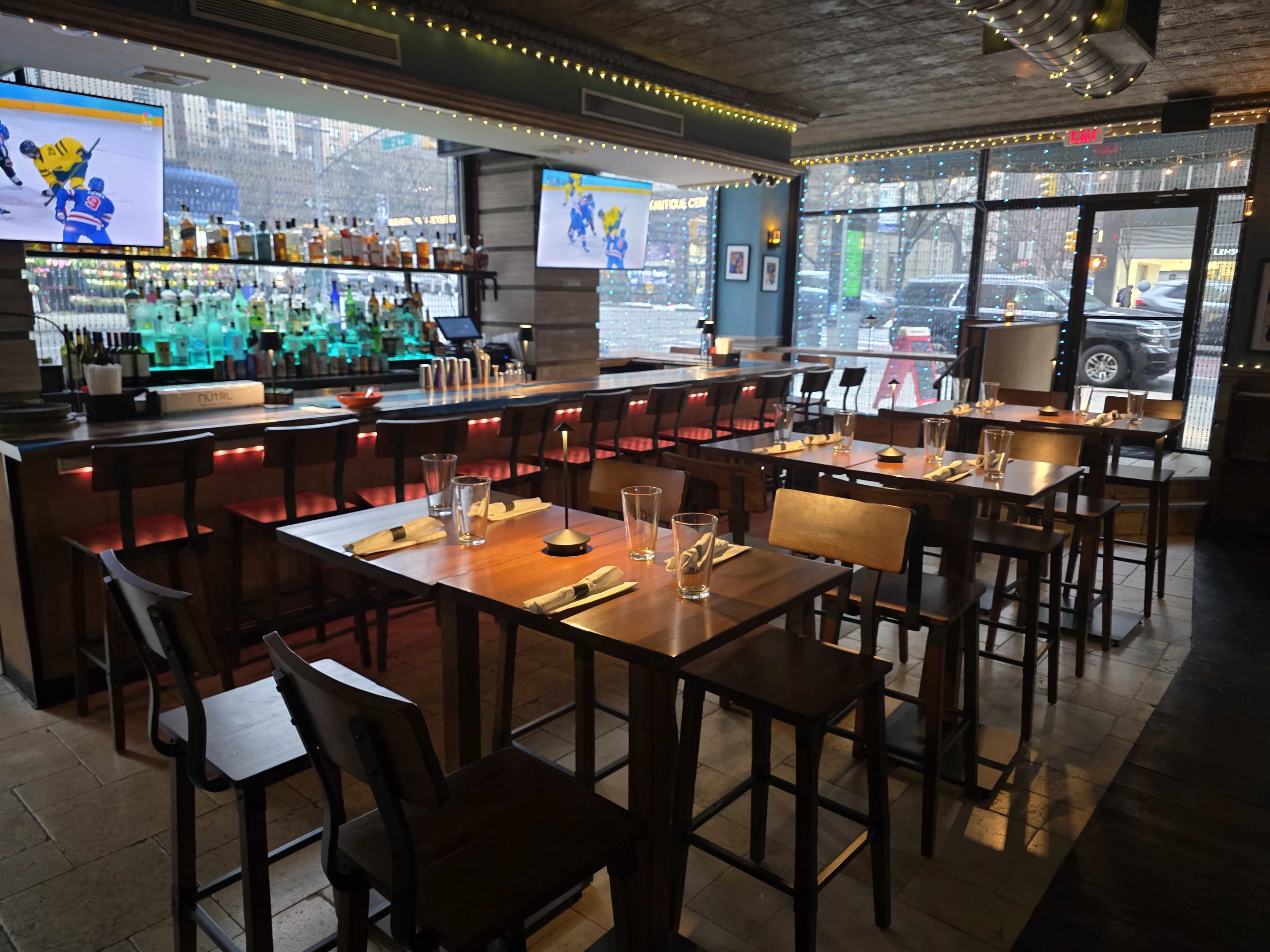 The image shows a modern restaurant interior with a bar featuring illuminated shelves, tall tables, and bar stools, while large windows provide views of the street outside.