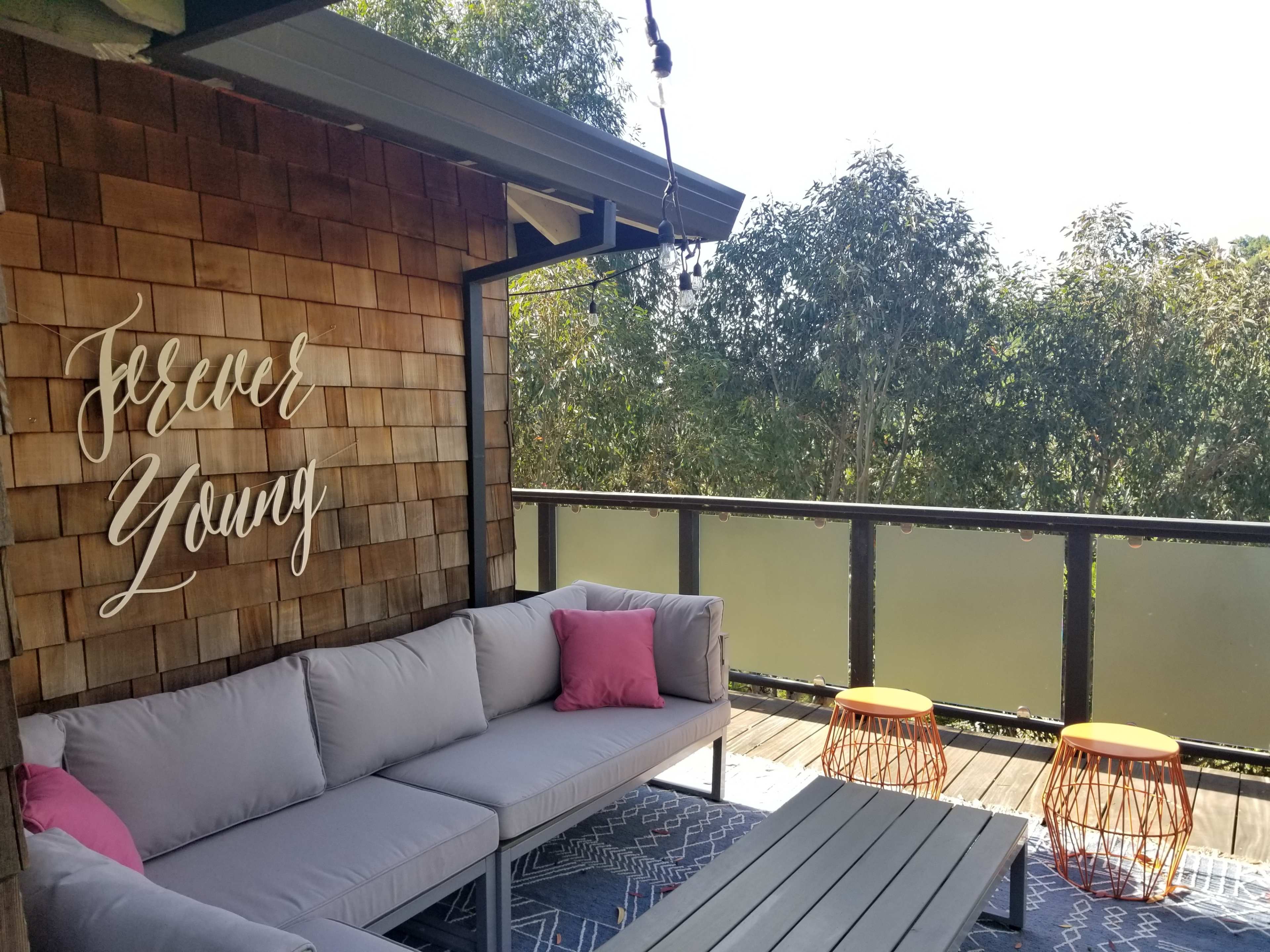 A cozy outdoor seating area features a gray sofa with pink cushions and two orange tables, against a backdrop of greenery and a wooden wall displaying the words "Forever Young."