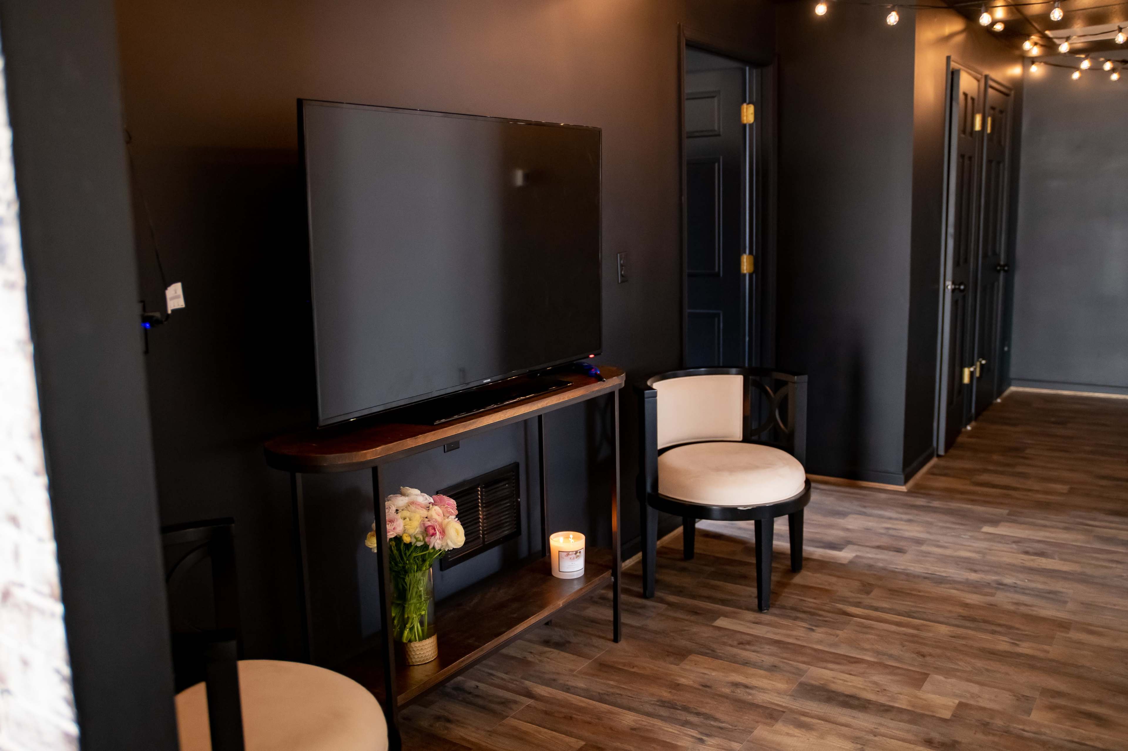 The image shows a dimly lit room with a large television mounted on a wall and a small table beneath it, flanked by two cushioned chairs.
