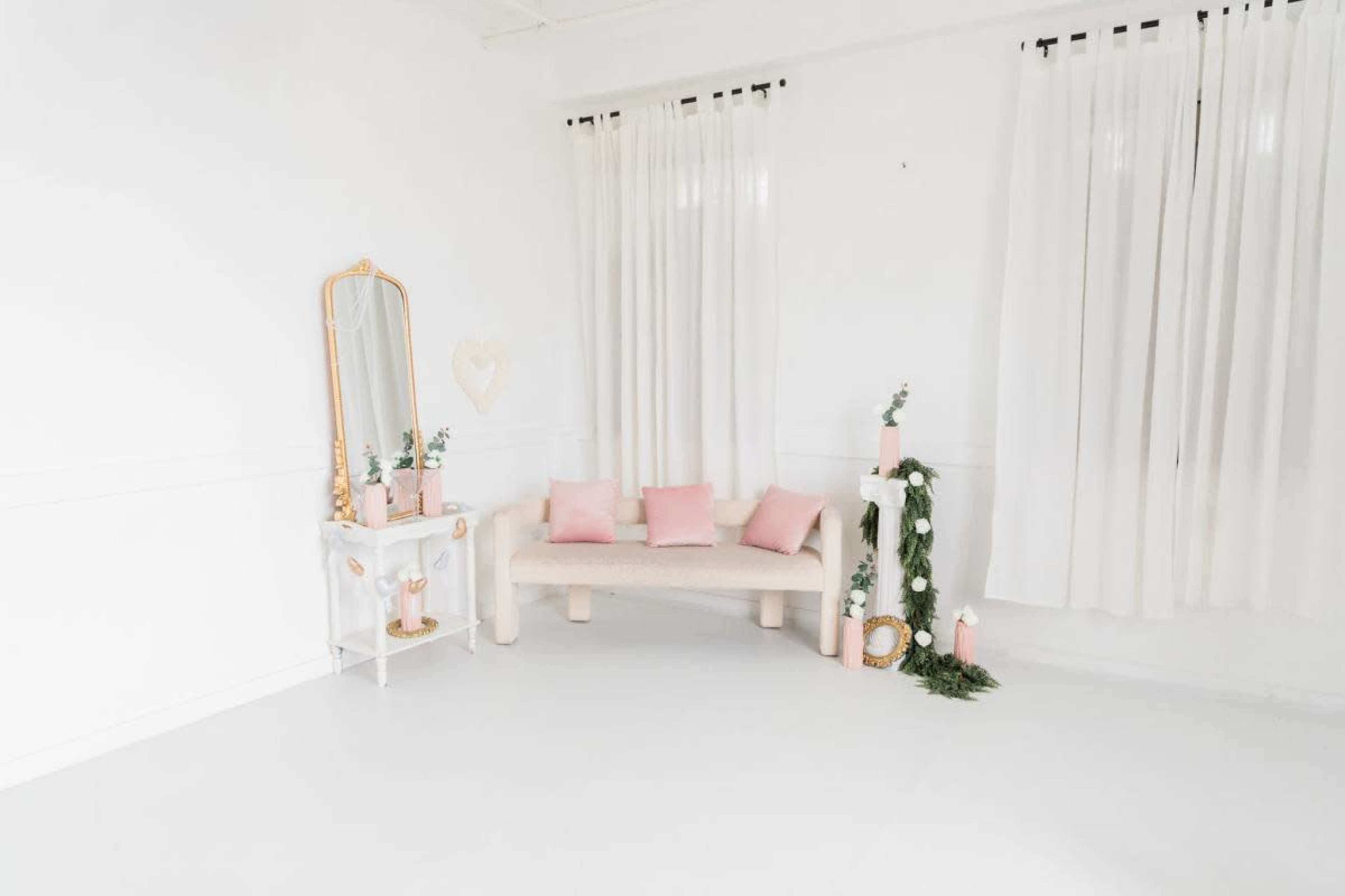 The image shows a bright, minimalist interior space featuring a pale sofa with pink cushions, a white table adorned with potted plants, and sheer white curtains.