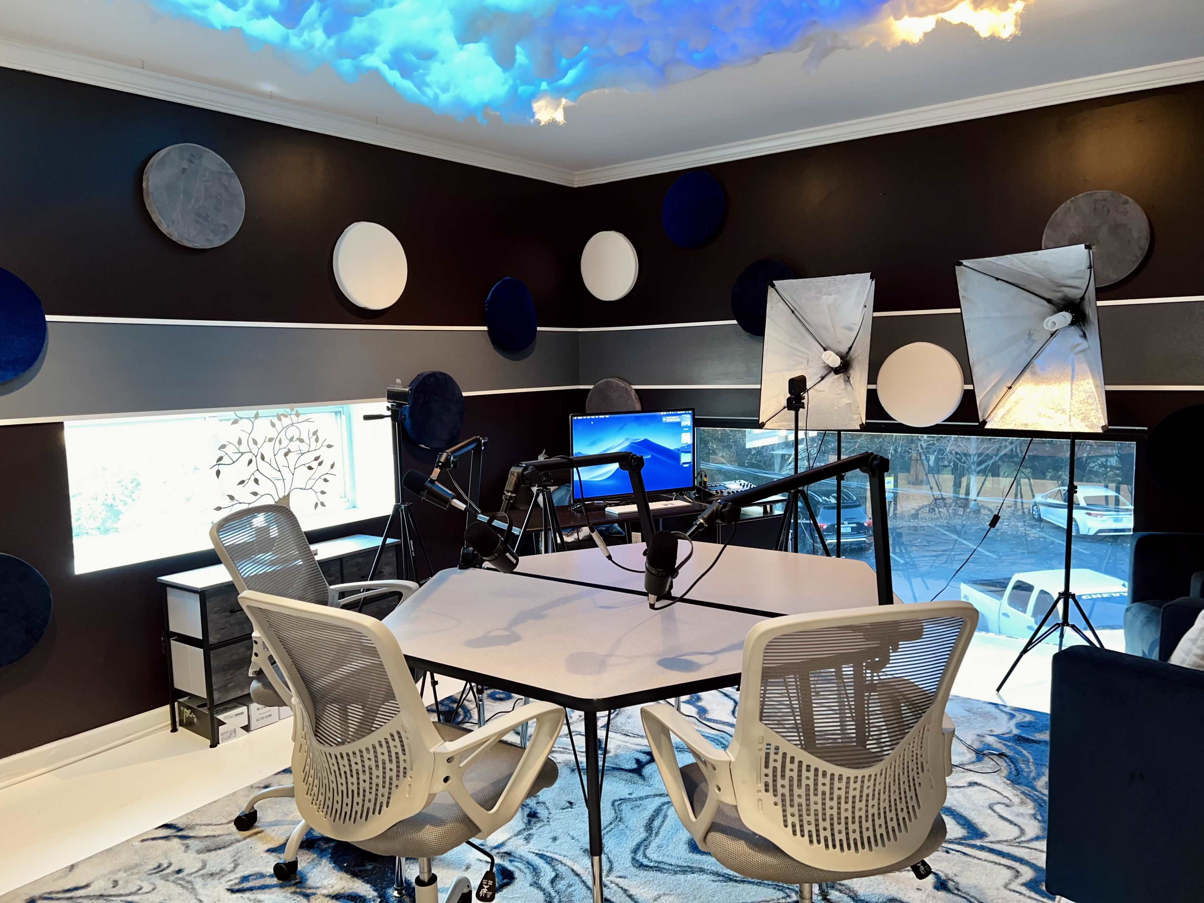 The image displays a modern podcast studio featuring a central table with microphones, white chairs, and soft lighting in a room accented with blue and white decor.