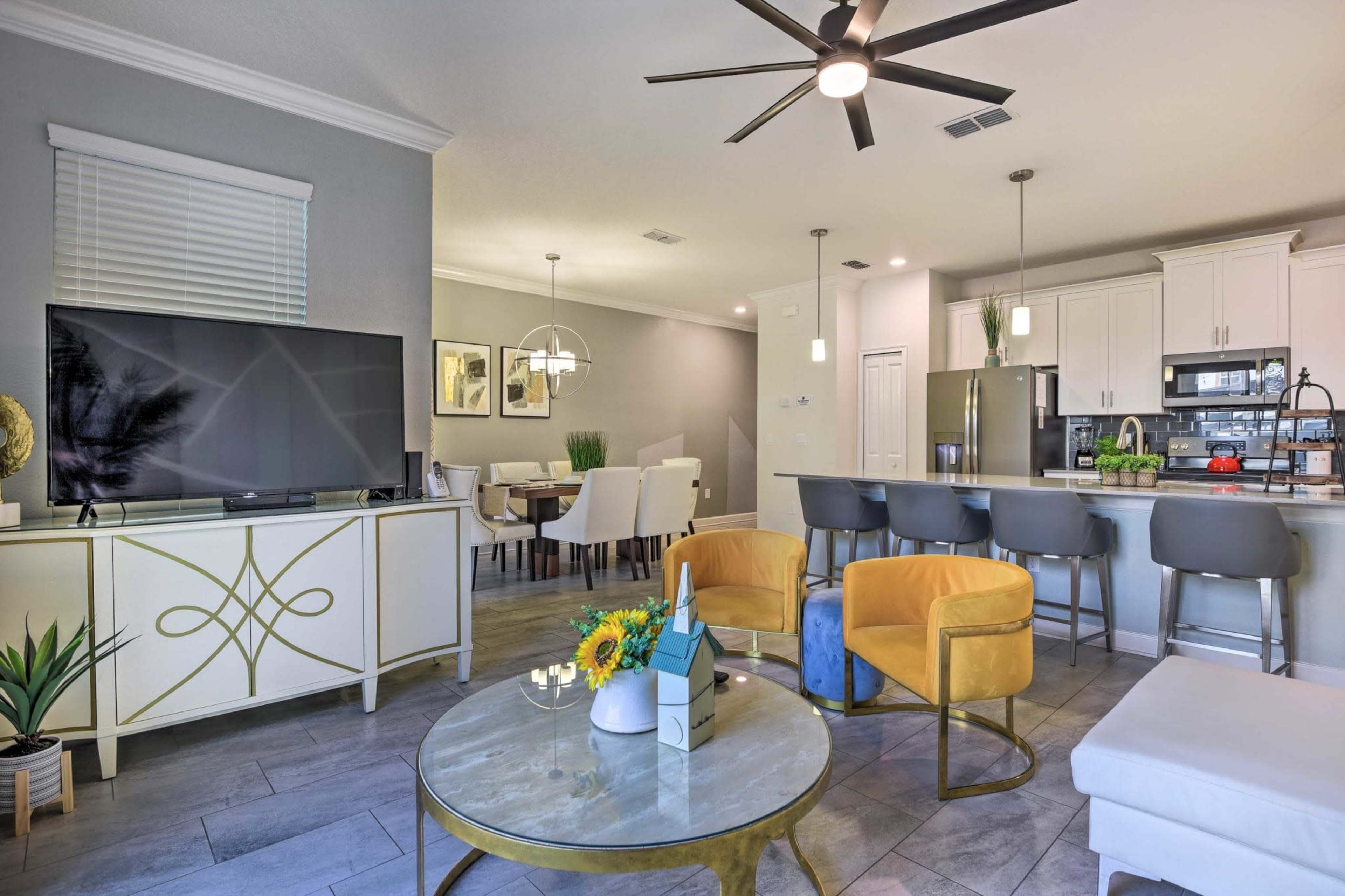 The image shows a modern living room with a flat-screen TV, yellow chairs, a coffee table, and a connected dining area featuring a kitchen countertop with bar stools.