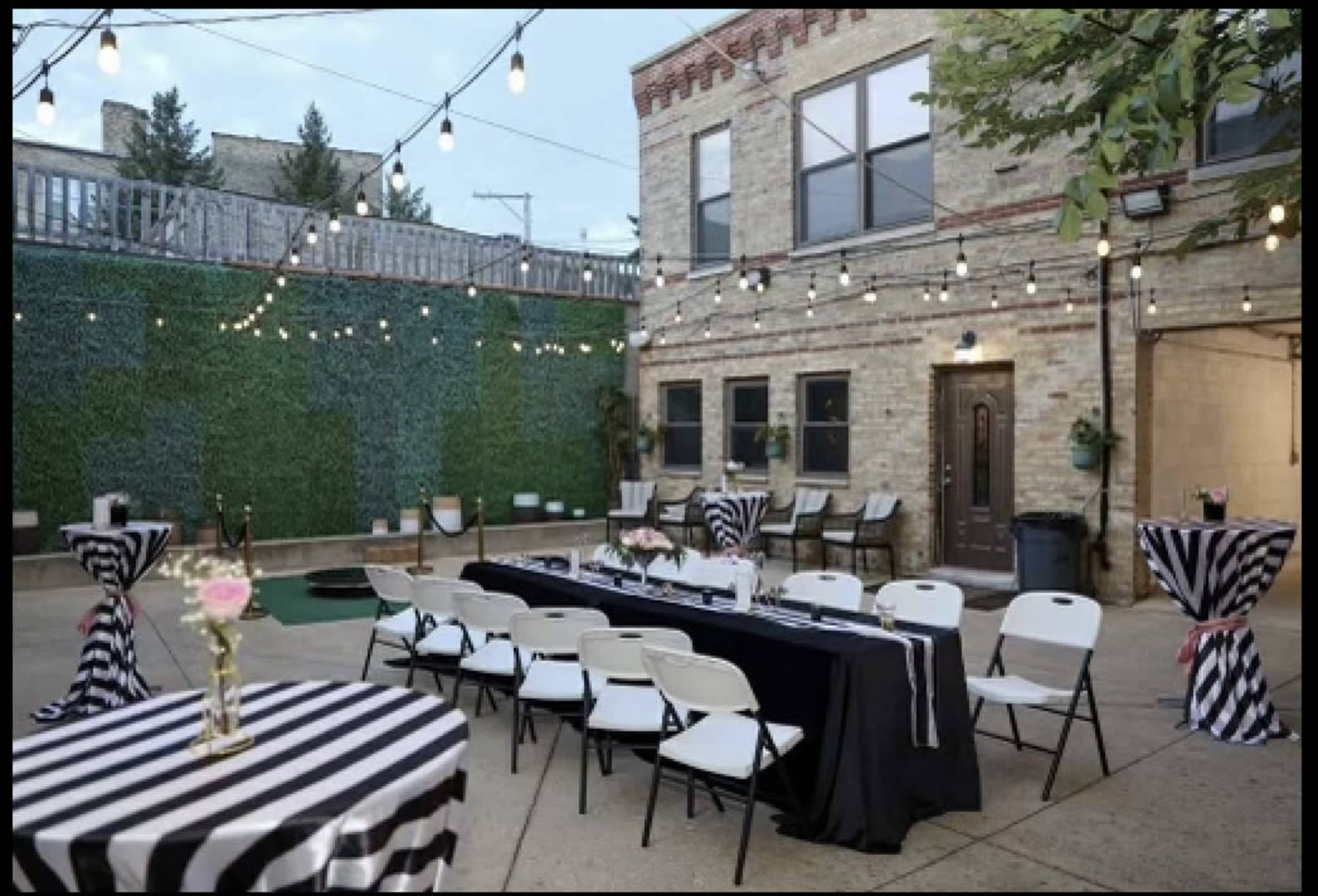 The image shows an outdoor event space with striped tablecloths, a long black table, chairs, and decorative string lights above, surrounded by greenery.