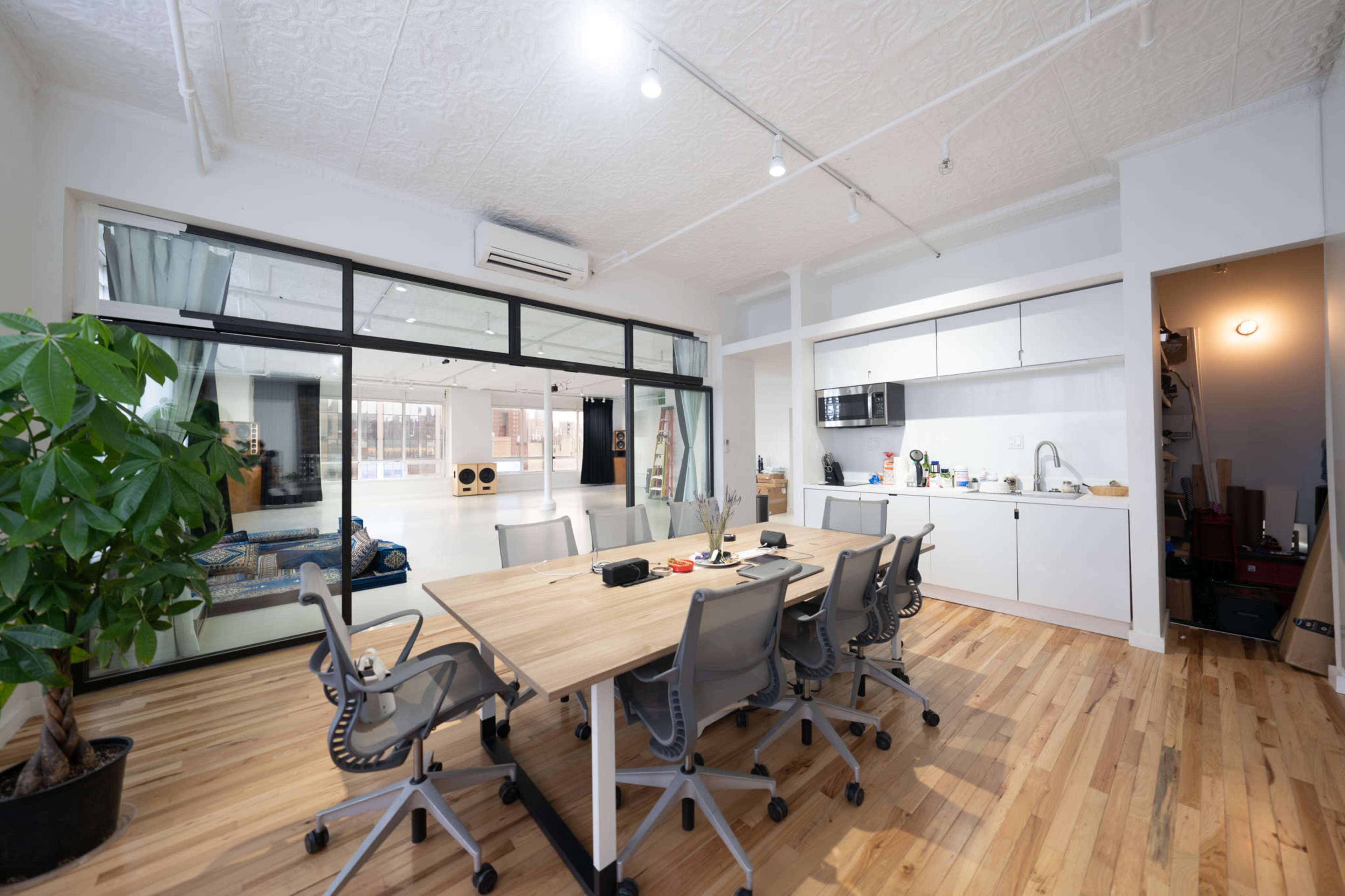 A spacious office features a large wooden table surrounded by ergonomic chairs, with a kitchenette and glass doors leading to a separate area.