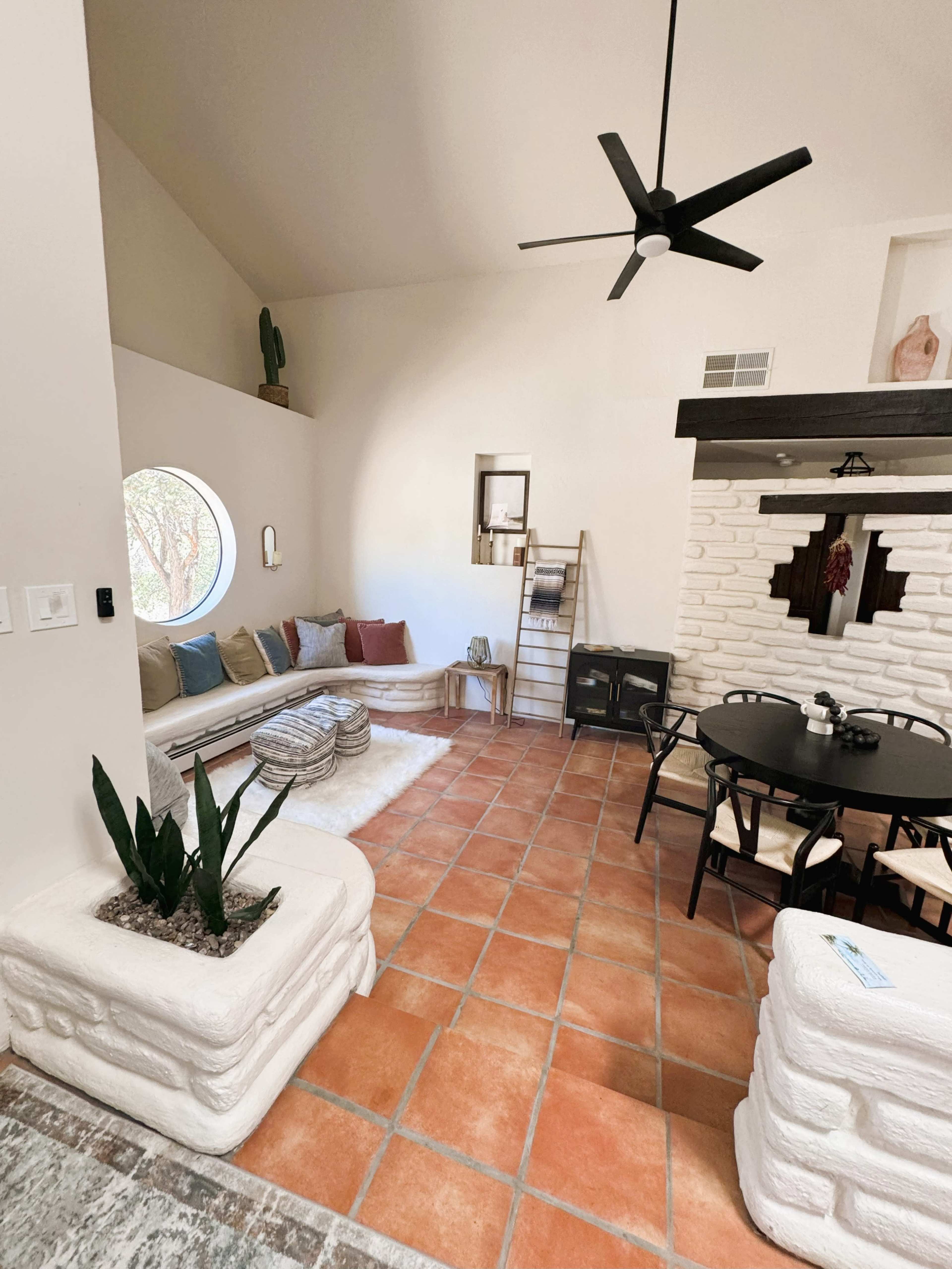 A modern living room with a circular window, a sofa arranged with colorful pillows, a dining table, and a decorative wall featuring a fireplace and potted plants.