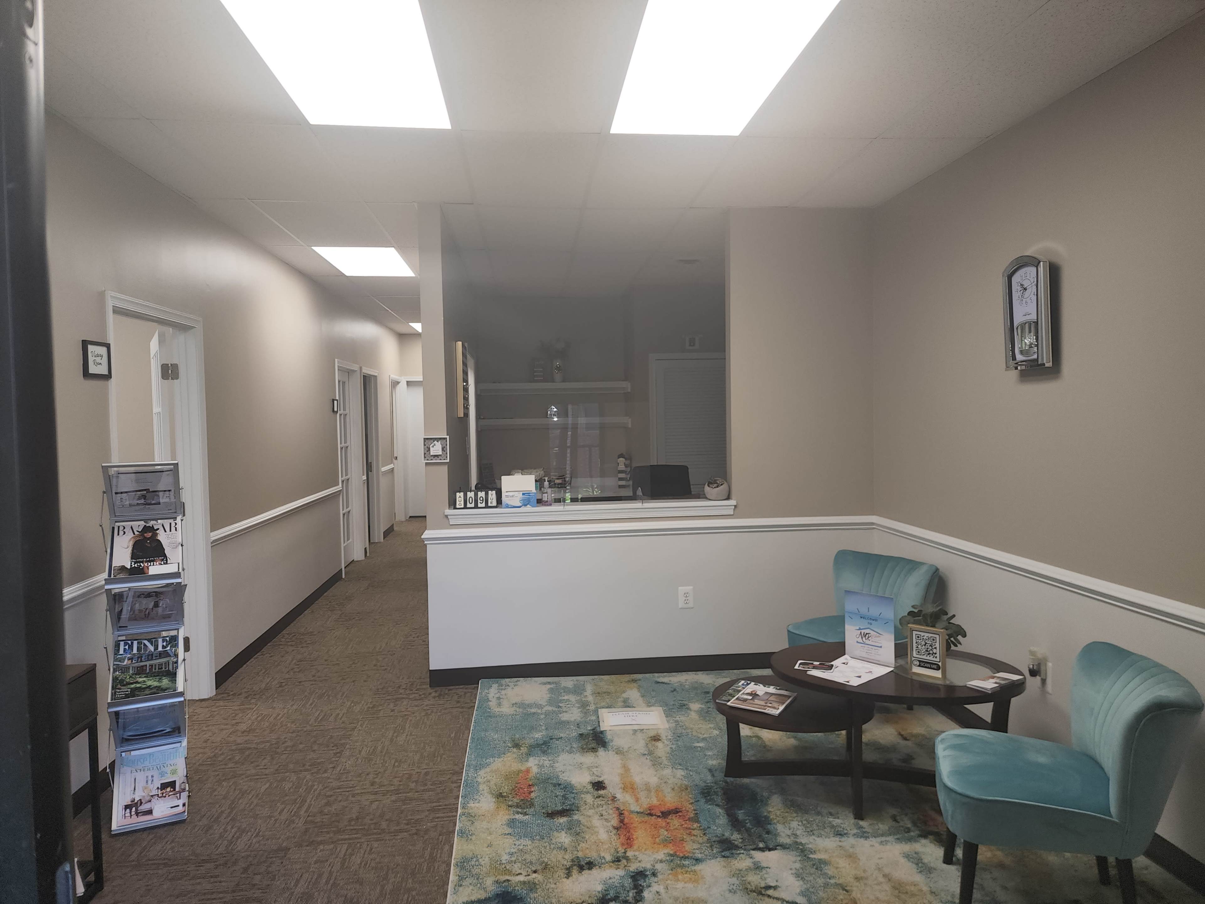 A waiting area with two blue chairs and a coffee table, positioned in front of a colorful rug, while a corridor with white doors leads to other rooms in the background.
