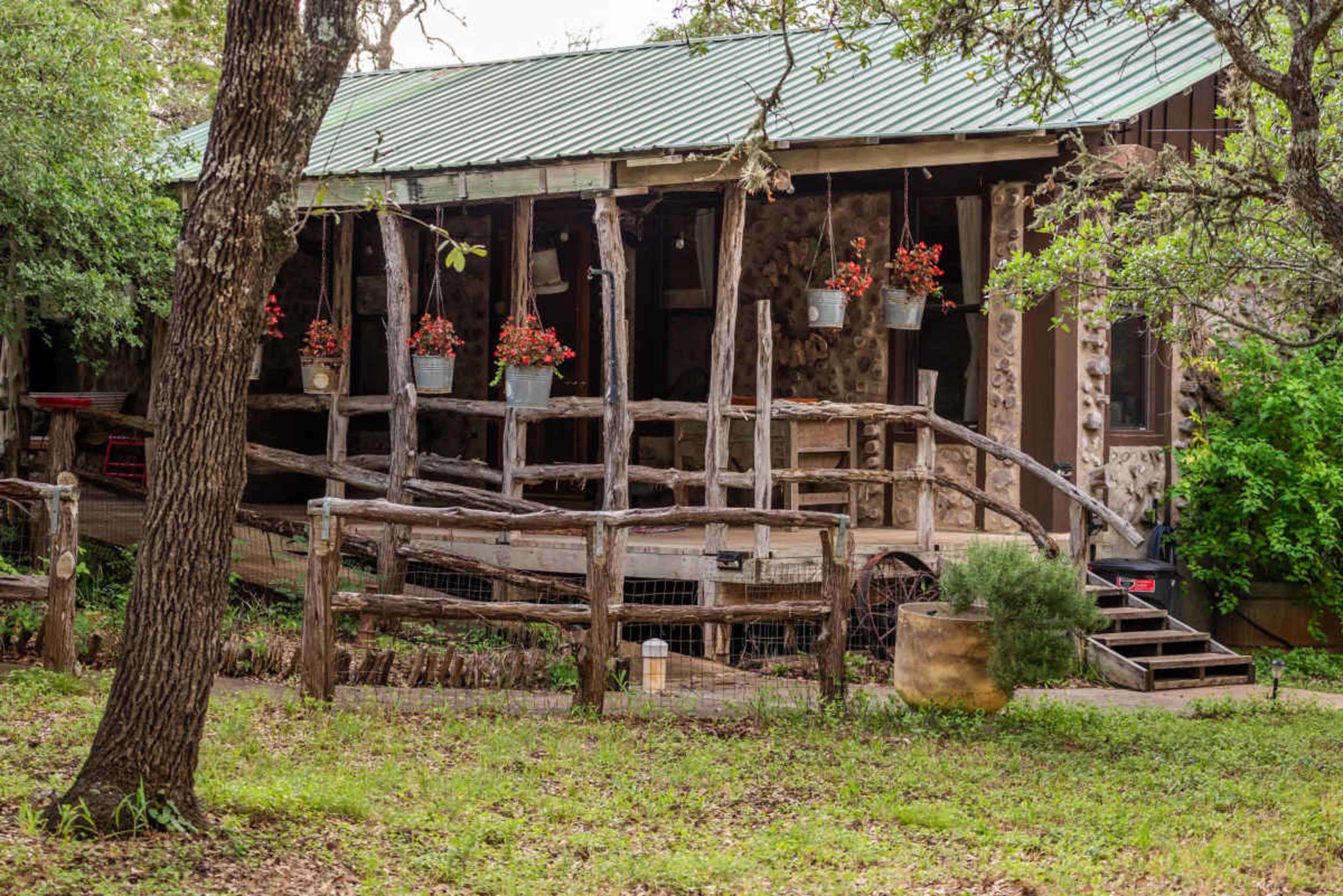 The image shows a rustic house with a green metal roof, featuring wooden supports and flower pots hanging from the porch.