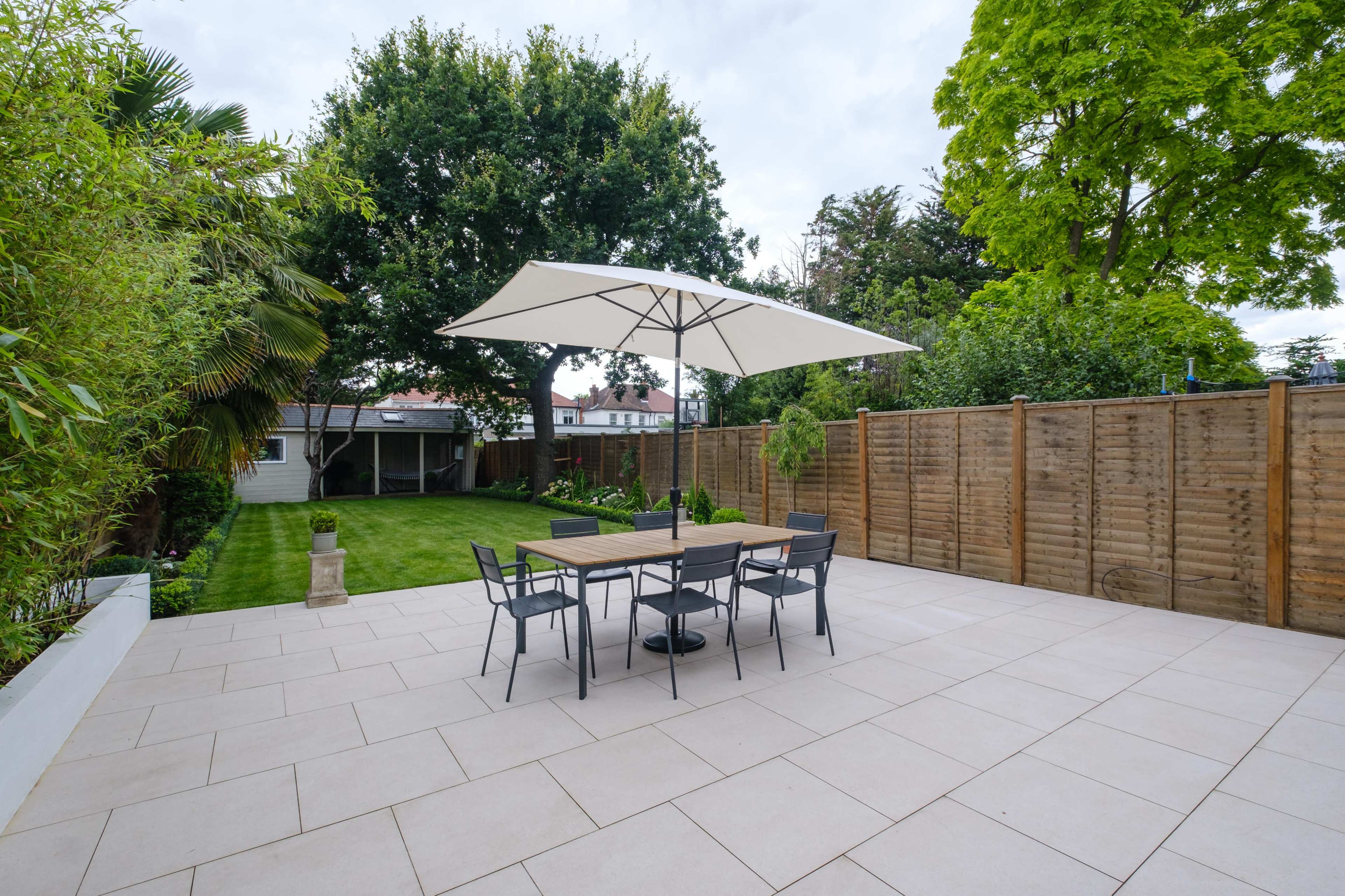 A patio area with a table and chairs covered by a large umbrella, surrounded by a grassy yard and wooden fencing.