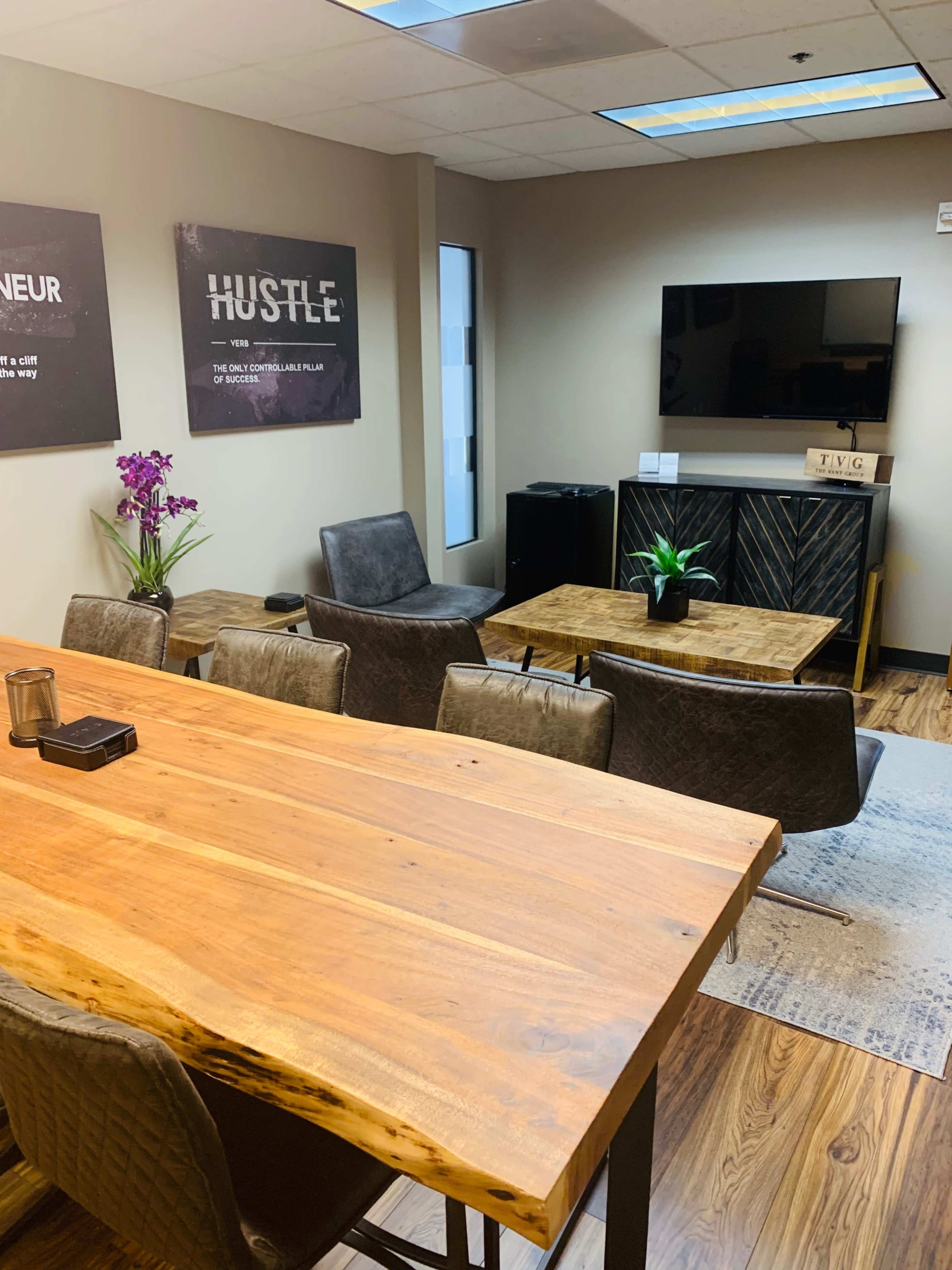 The image shows a modern office meeting space with a wooden table, leather chairs, a television mounted on the wall, and decorative plants.
