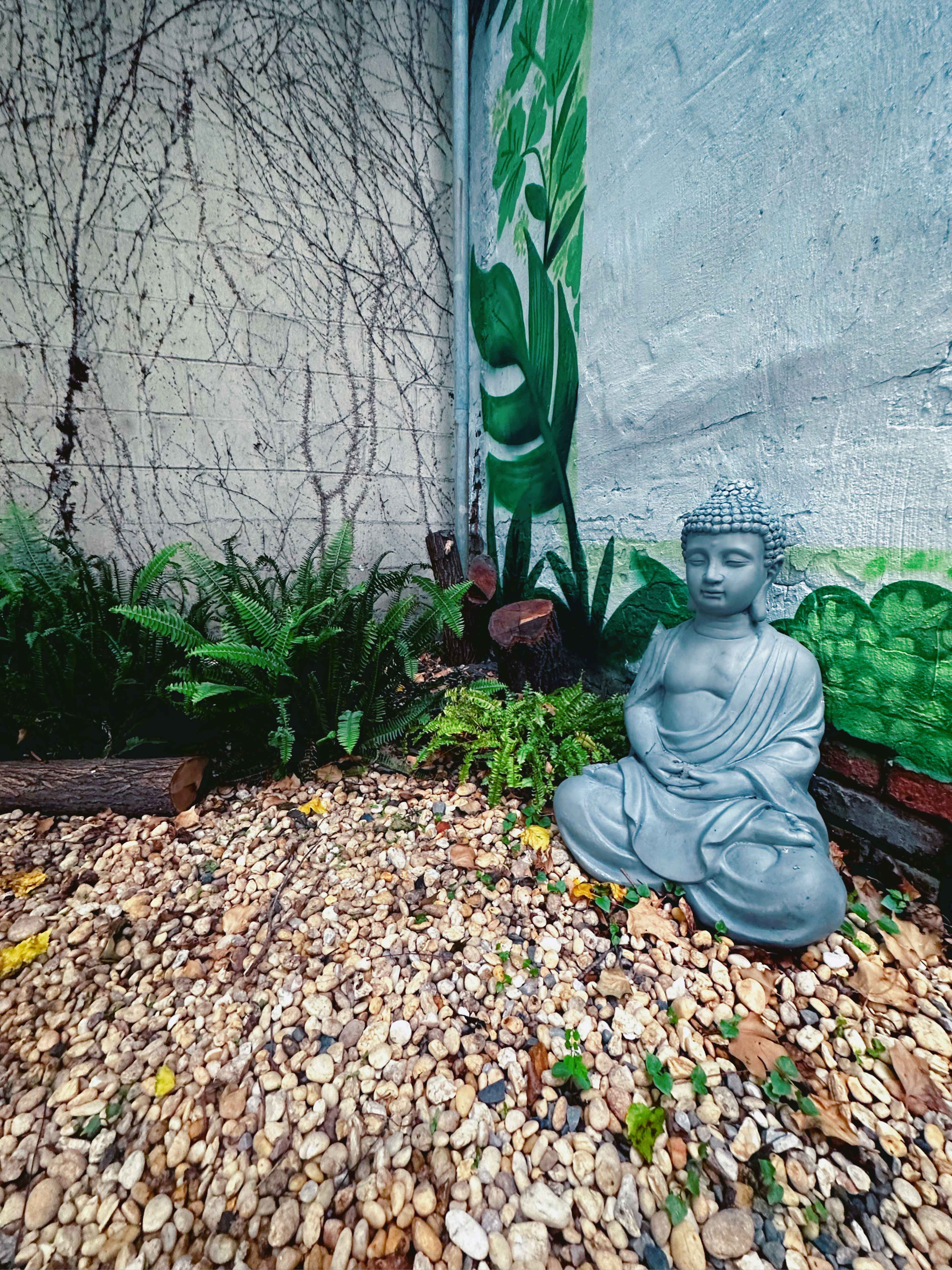 A Buddha statue sits among pebbles and ferns in a garden next to a painted wall of green leaves.