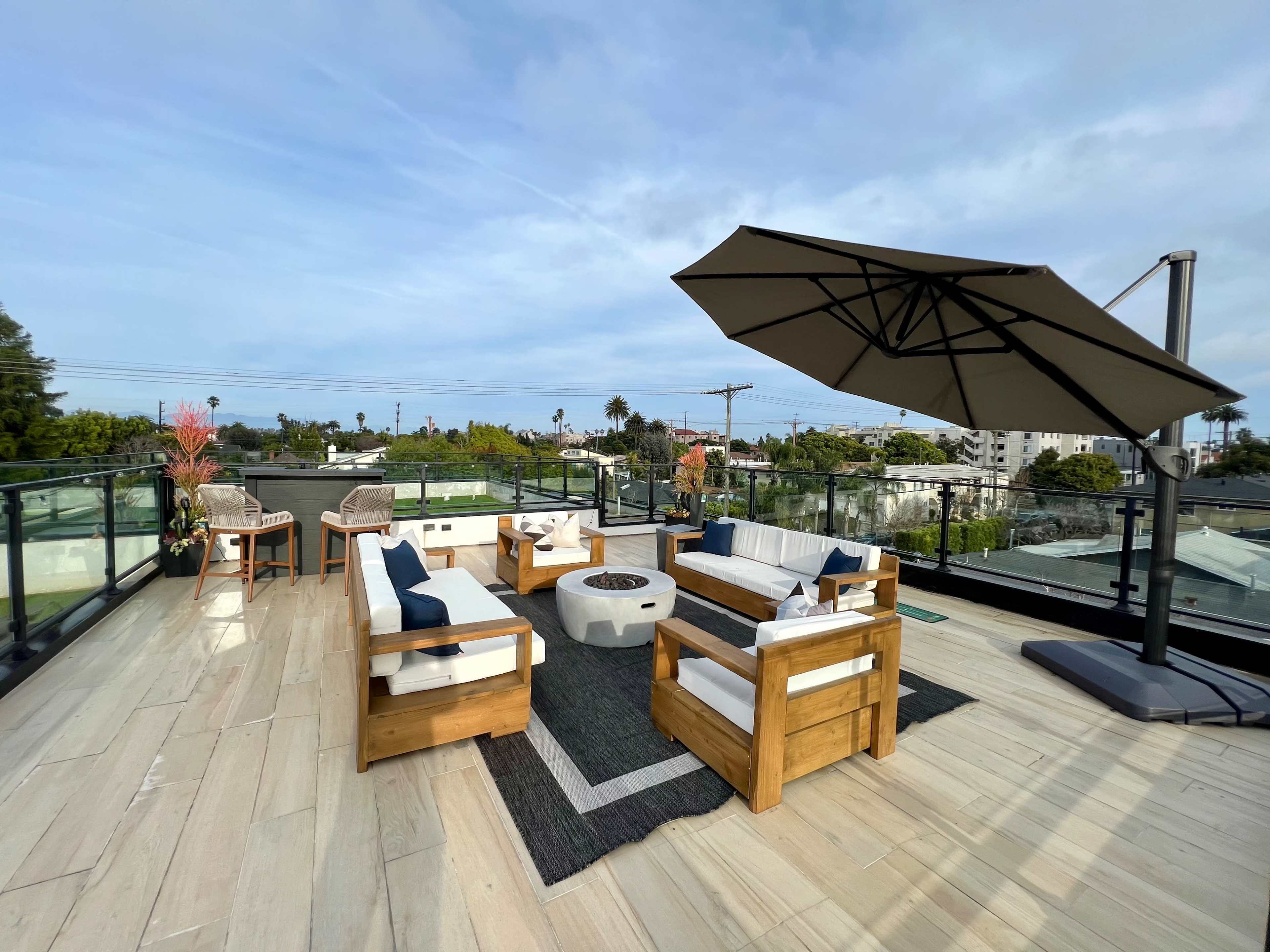 A spacious rooftop patio with wooden seating, a fire pit, and a large umbrella, surrounded by greenery and buildings.