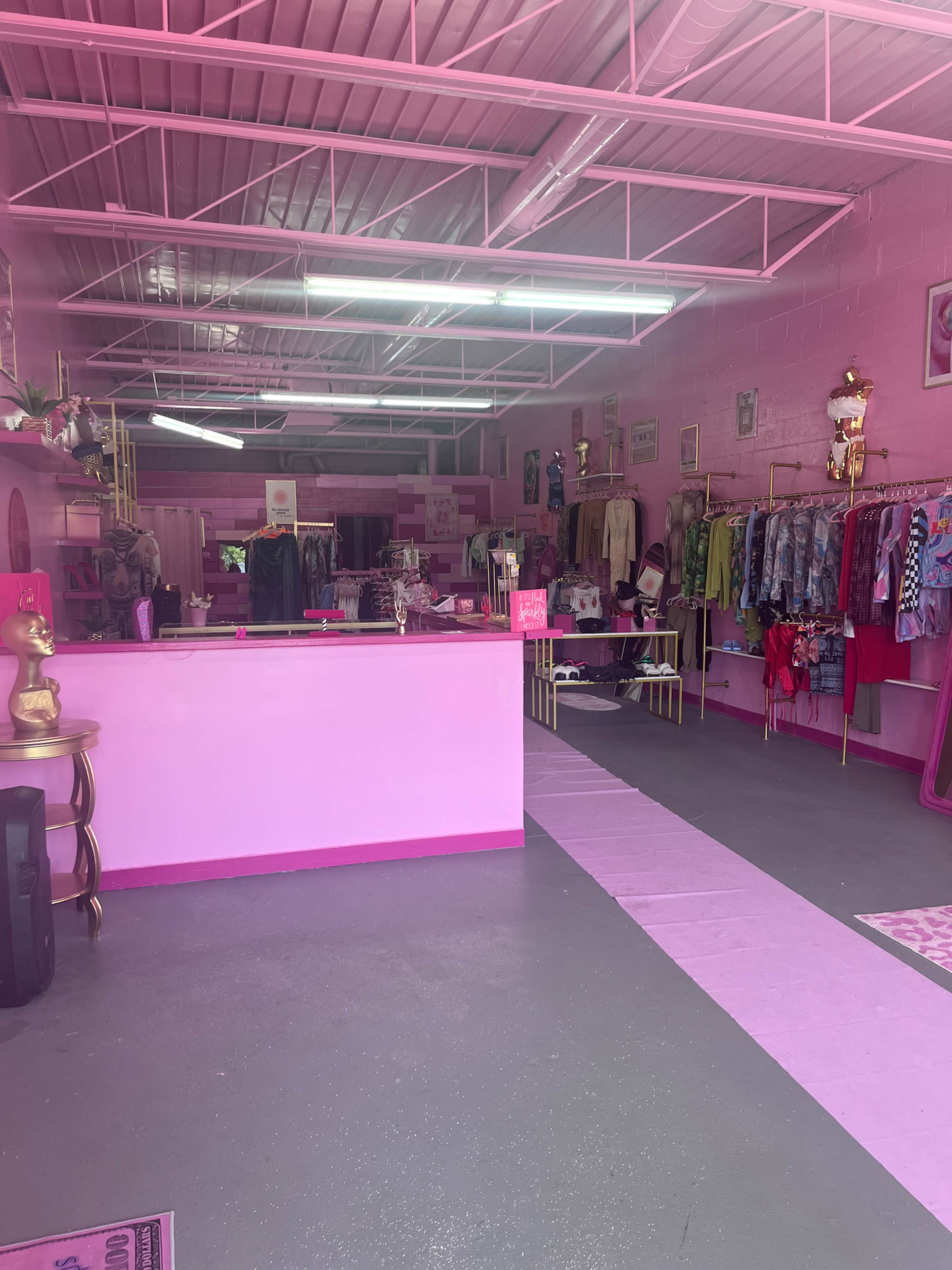 The image shows a brightly colored pink boutique featuring various clothing items on racks and decorations throughout the shop.