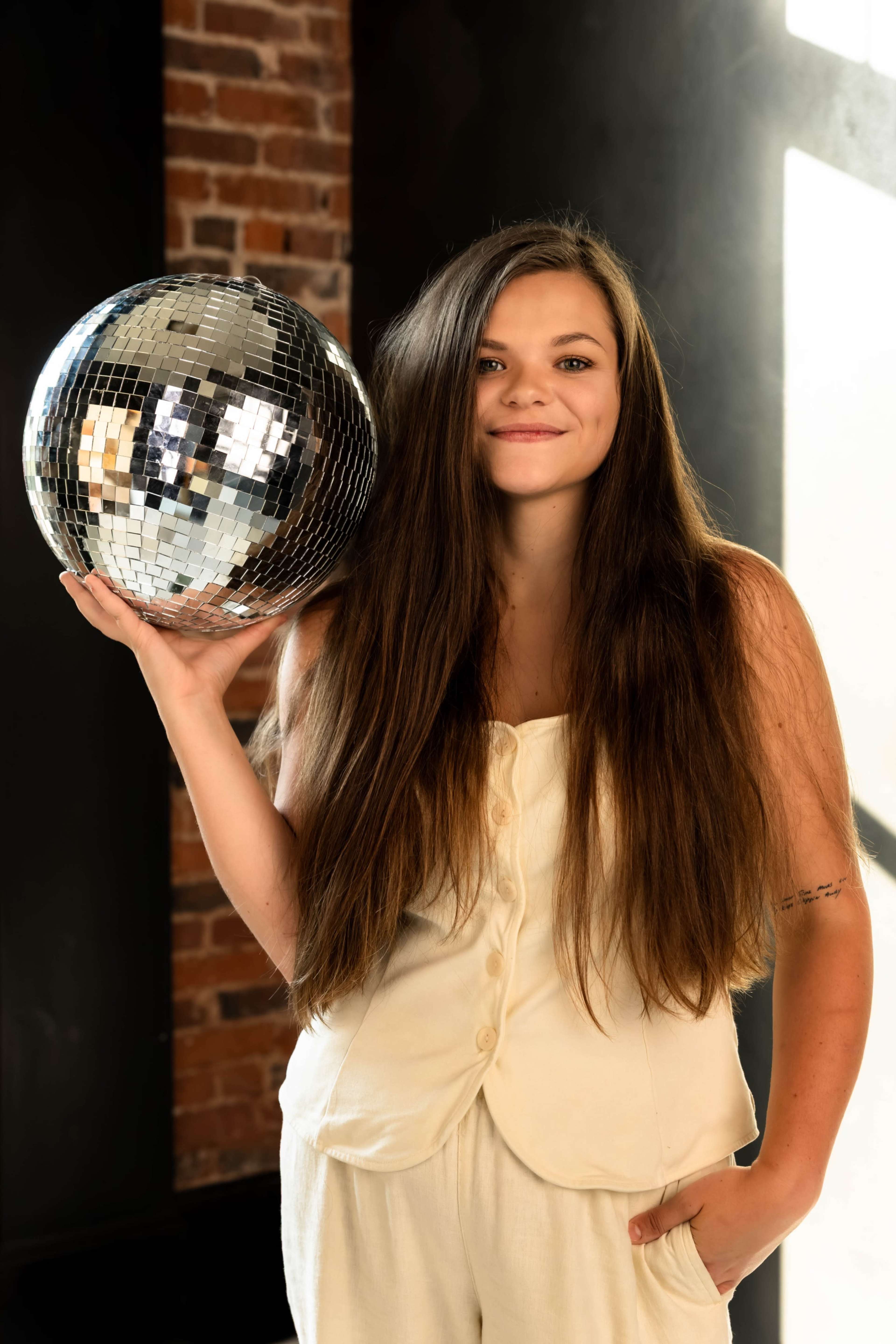 A person with long hair holds a disco ball in a brightly lit room with exposed brick walls.