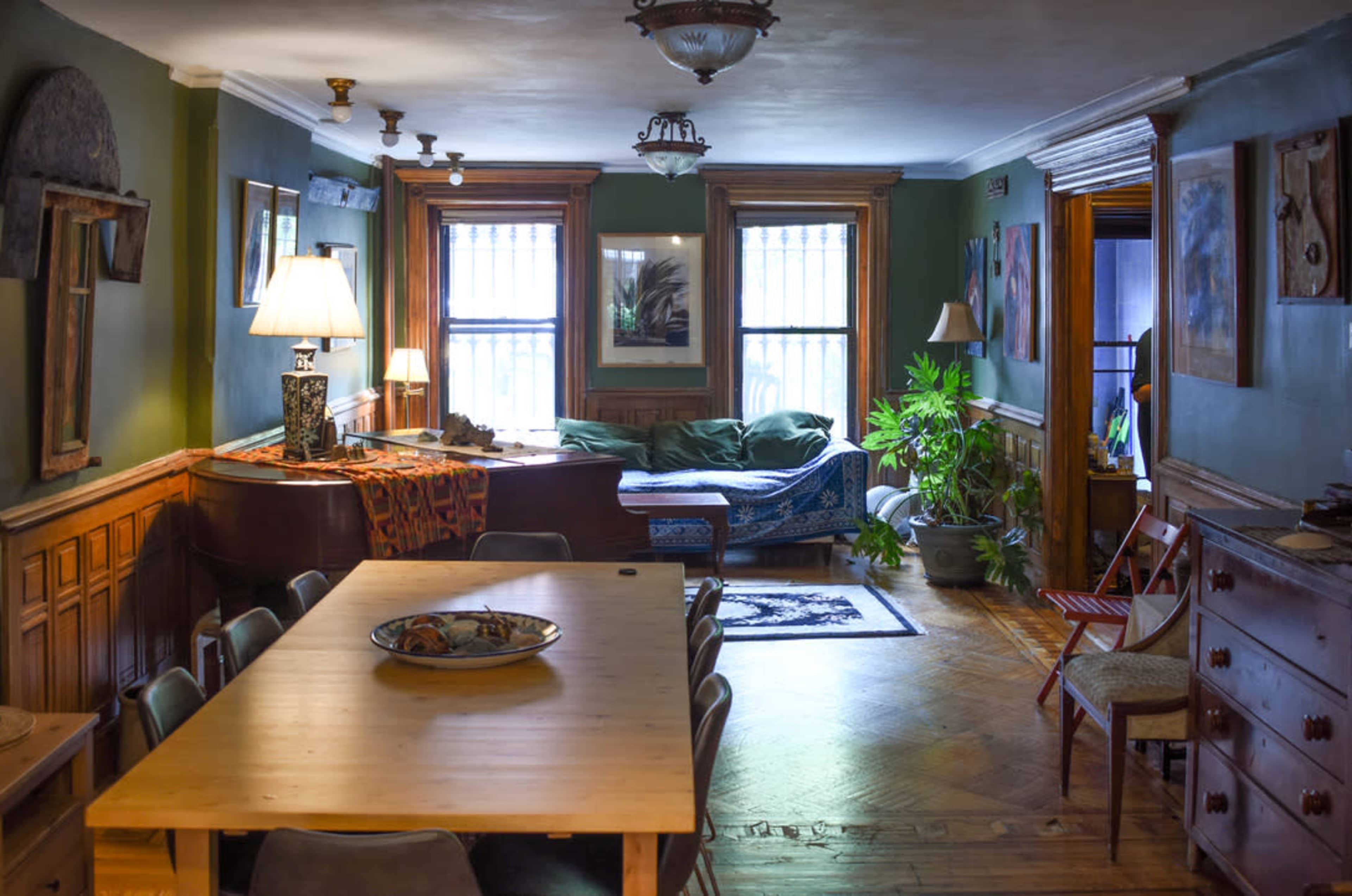A cozy living space with a wooden dining table, a grand piano, a green couch, and various plants, illuminated by natural light from windows.
