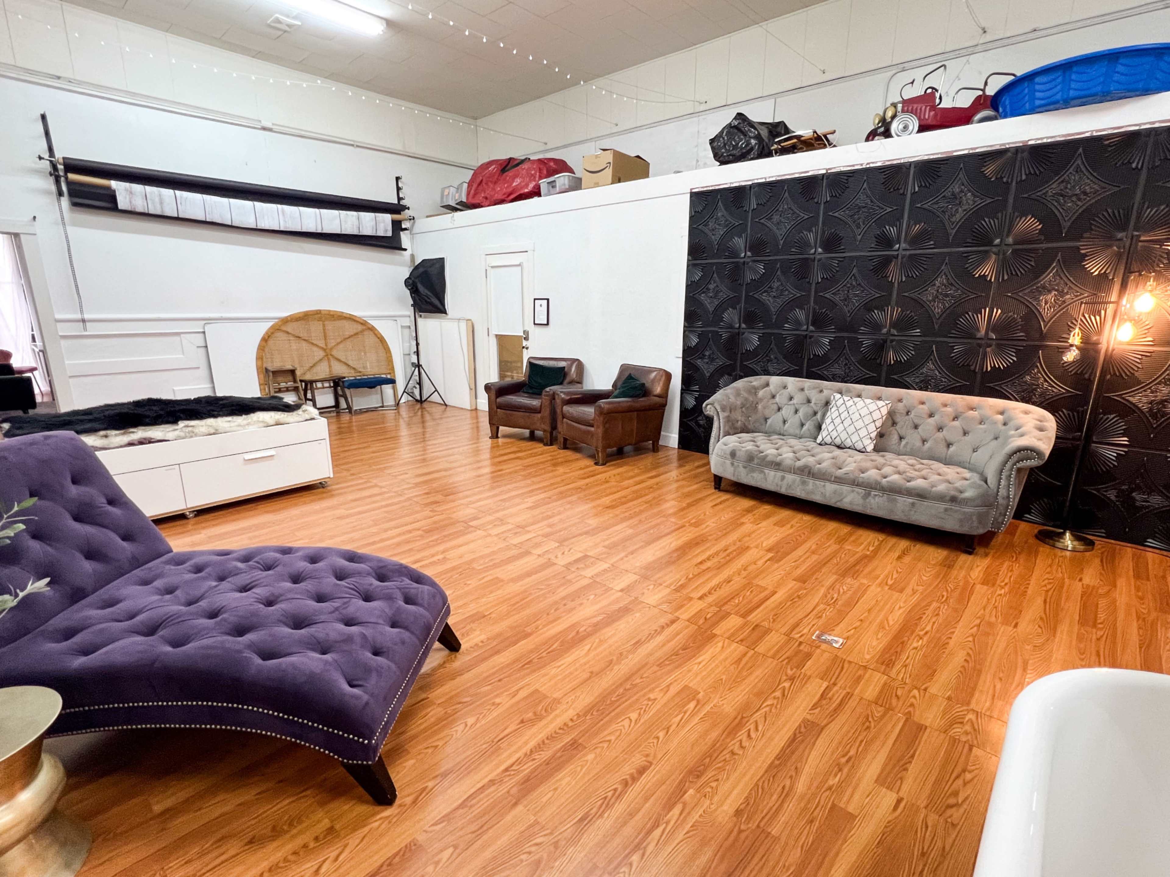 The room features a wooden floor, a gray tufted sofa, two brown armchairs, and various items stored along the walls, including a large decorative panel and a round backdrop.