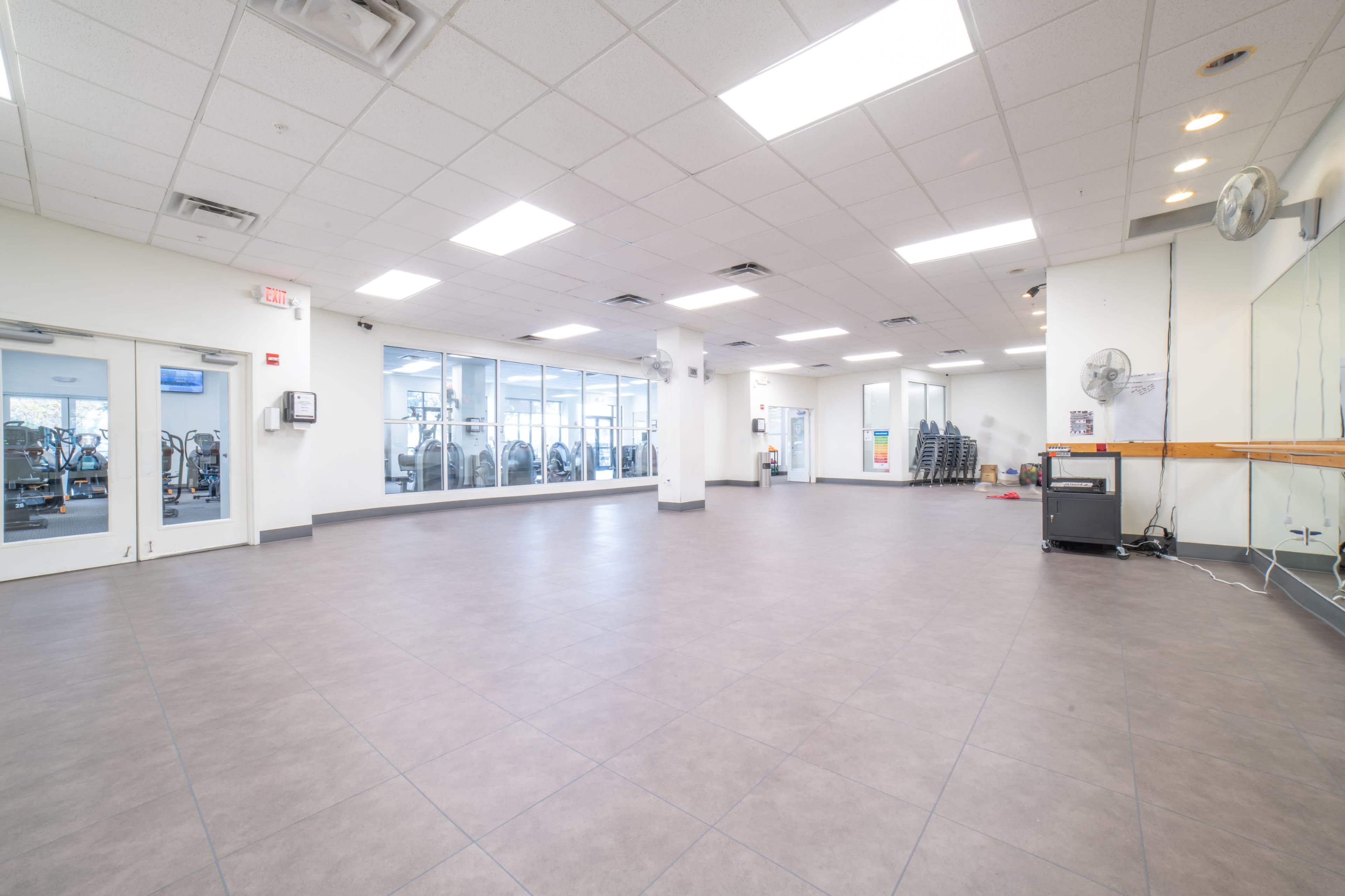 The image shows an empty fitness studio with tiled flooring, large windows, and gym equipment visible in the background.