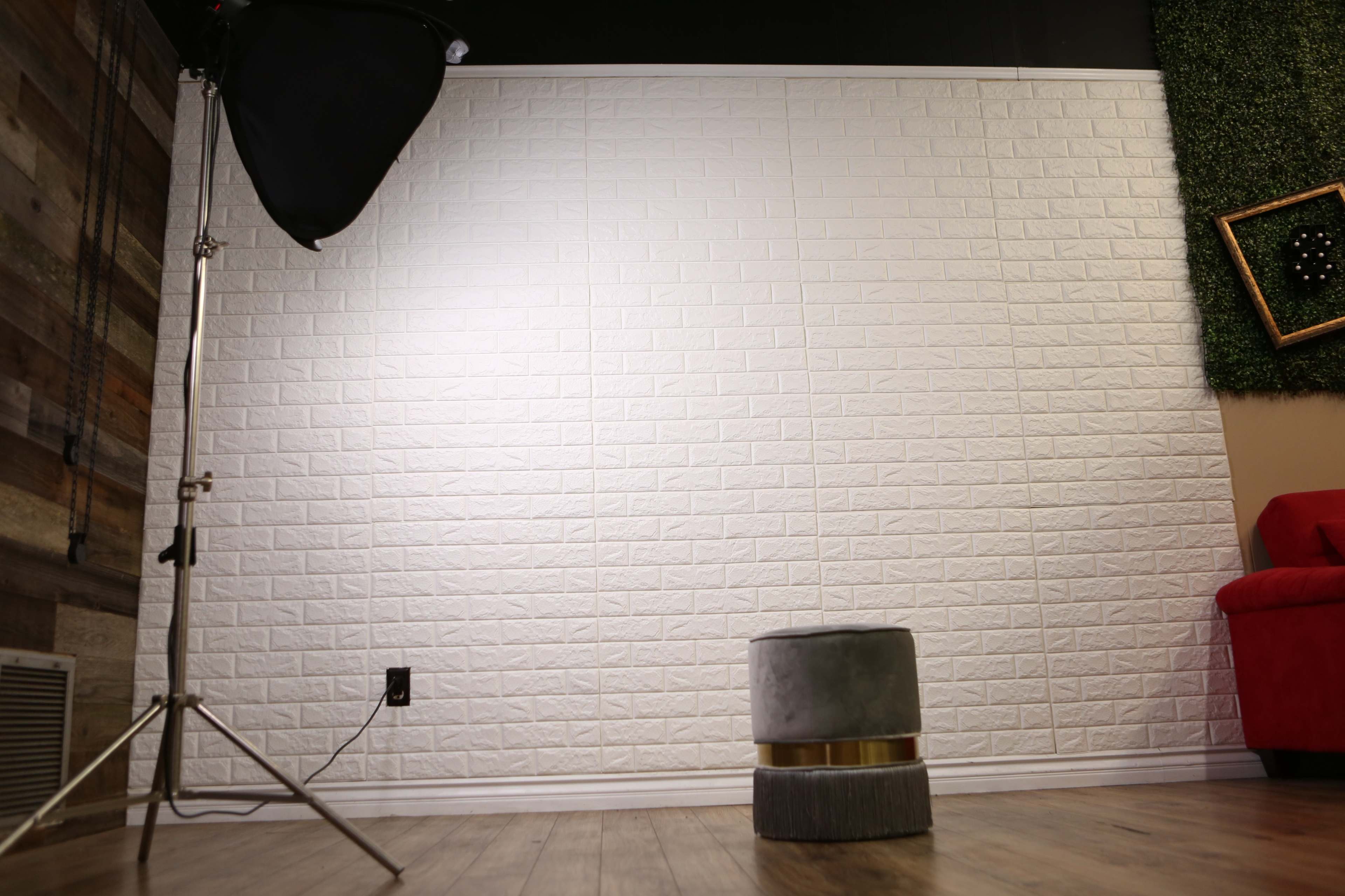 A spacious room with a white textured wall, a single gray stool, a studio light on a stand, and a red couch in the corner.
