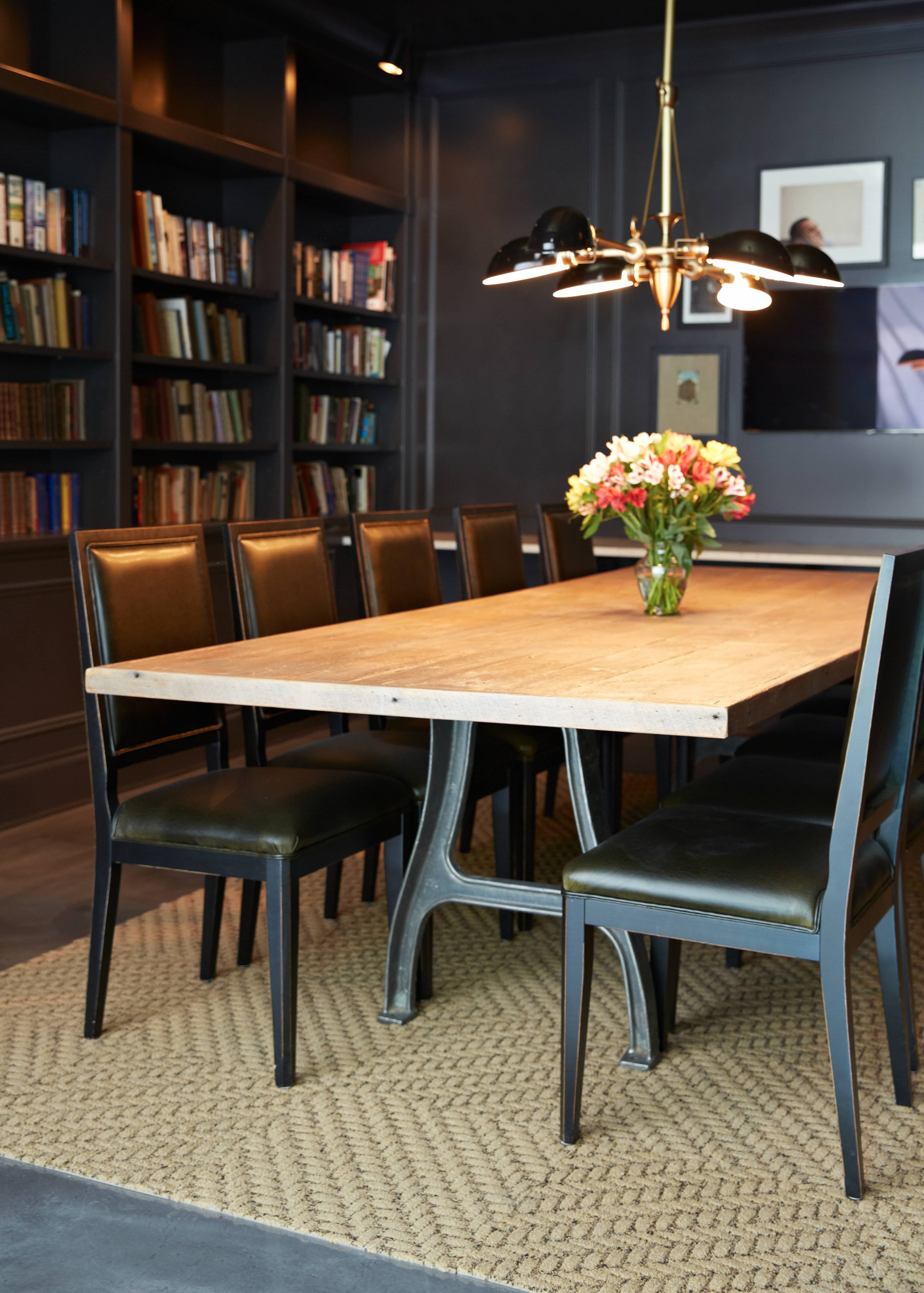 A long wooden dining table with black chairs is placed on a woven rug, surrounded by bookshelves filled with books and a flower vase at the center.