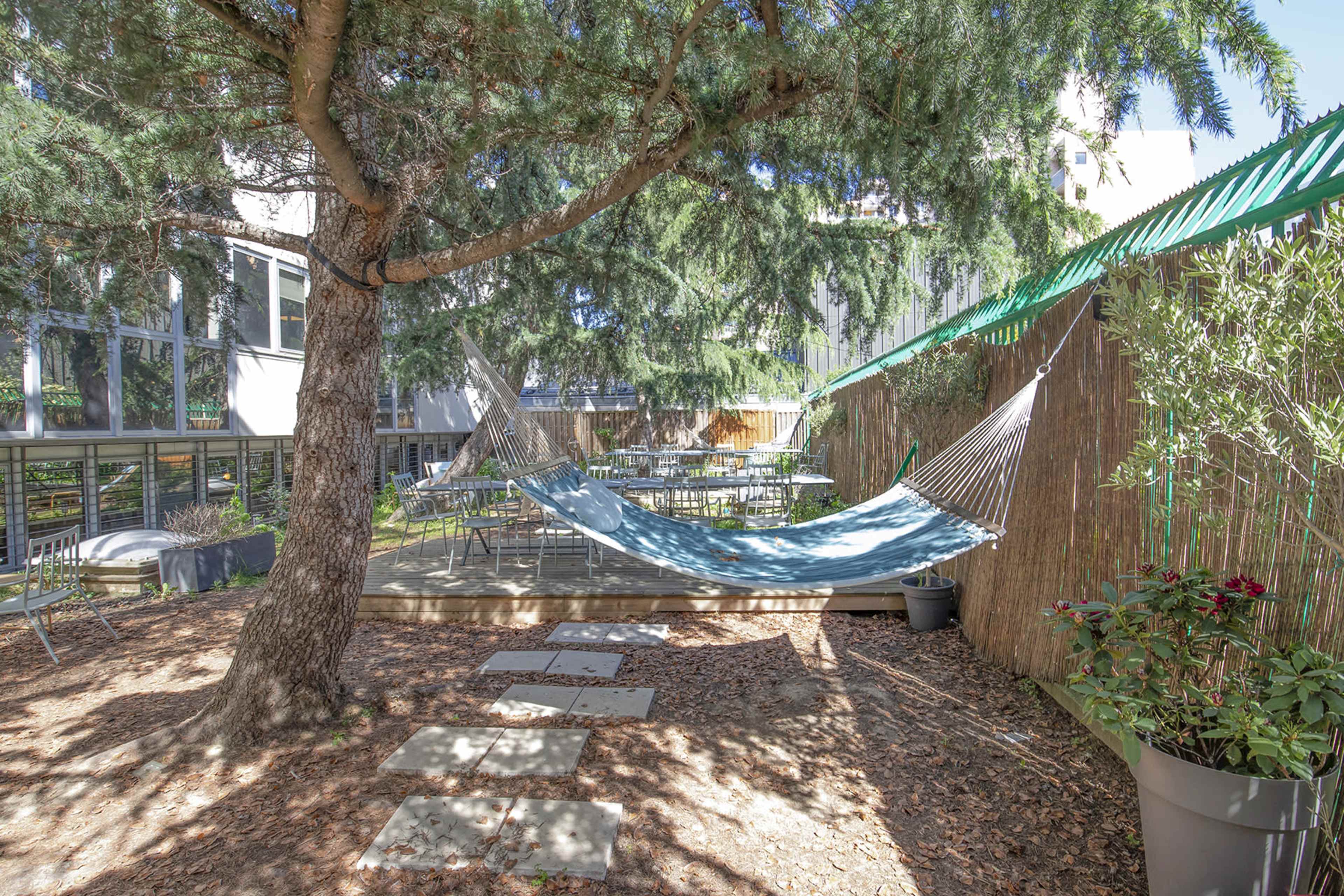 A hammock is suspended between two trees in a garden area surrounded by tables and greenery.