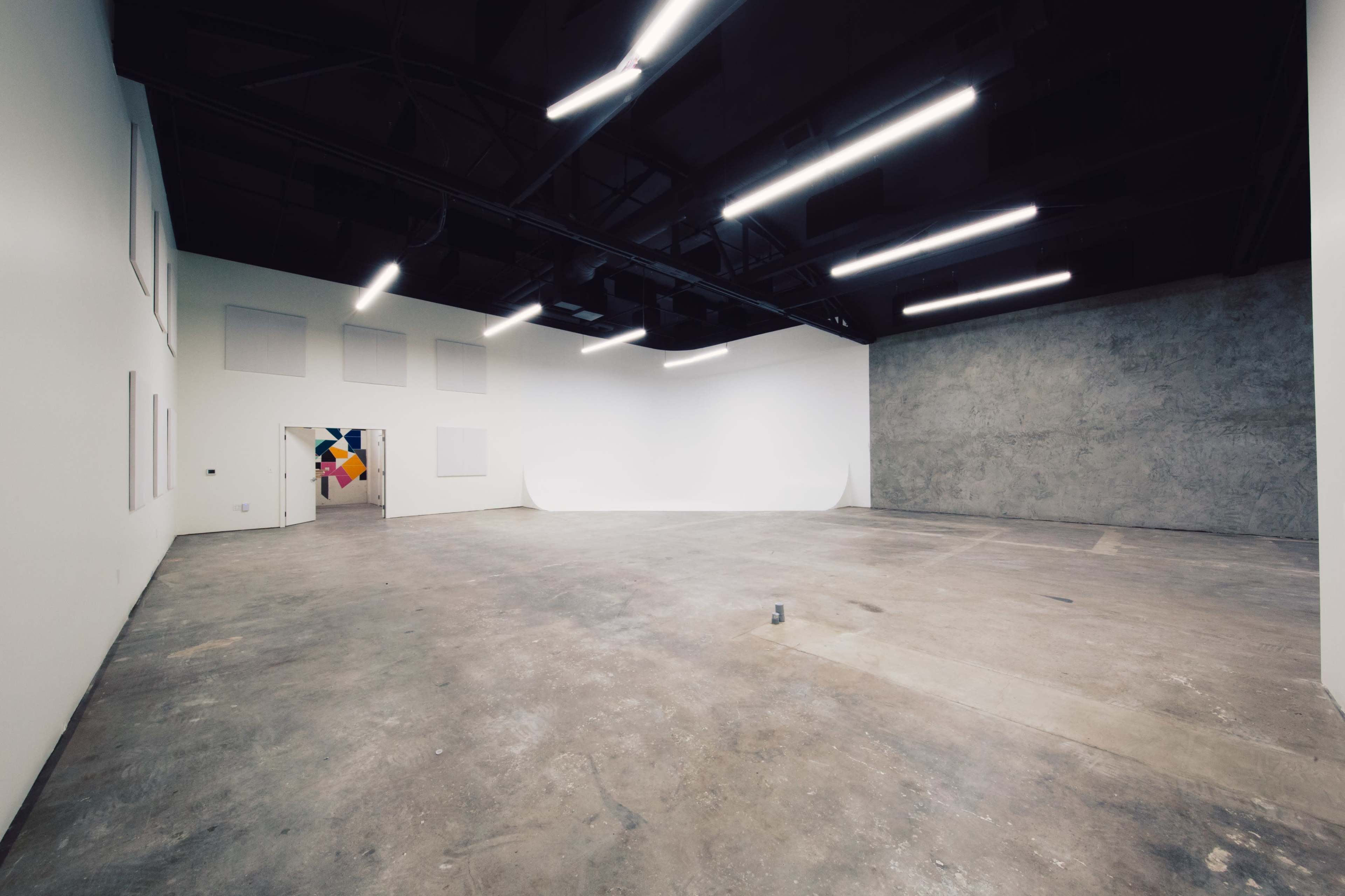 A spacious, empty gallery with white walls, a concrete wall on one side, and overhead fluorescent lighting.