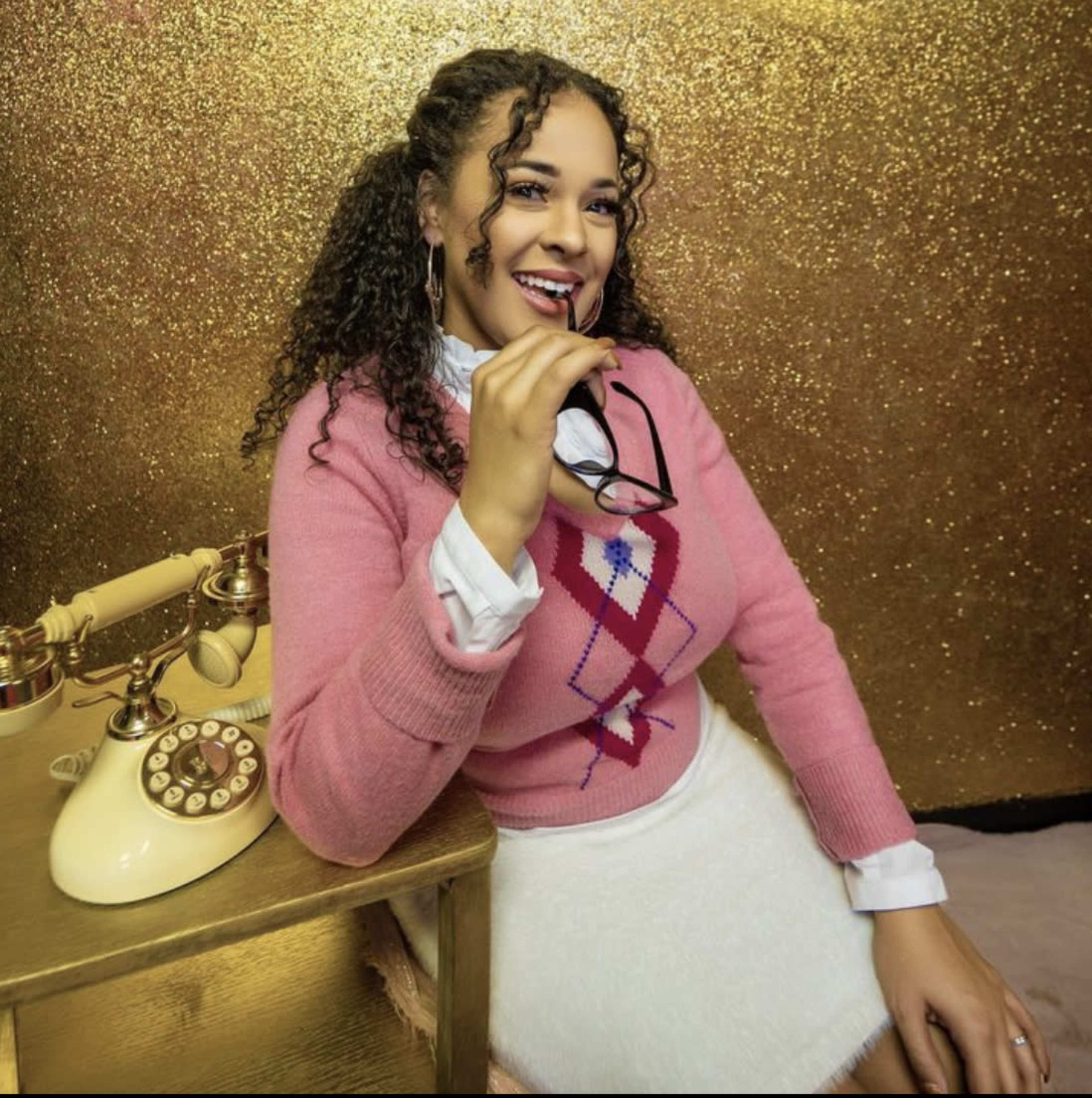 A woman with curly hair sits next to a vintage telephone, smiling while holding glasses and wearing a pink sweater.