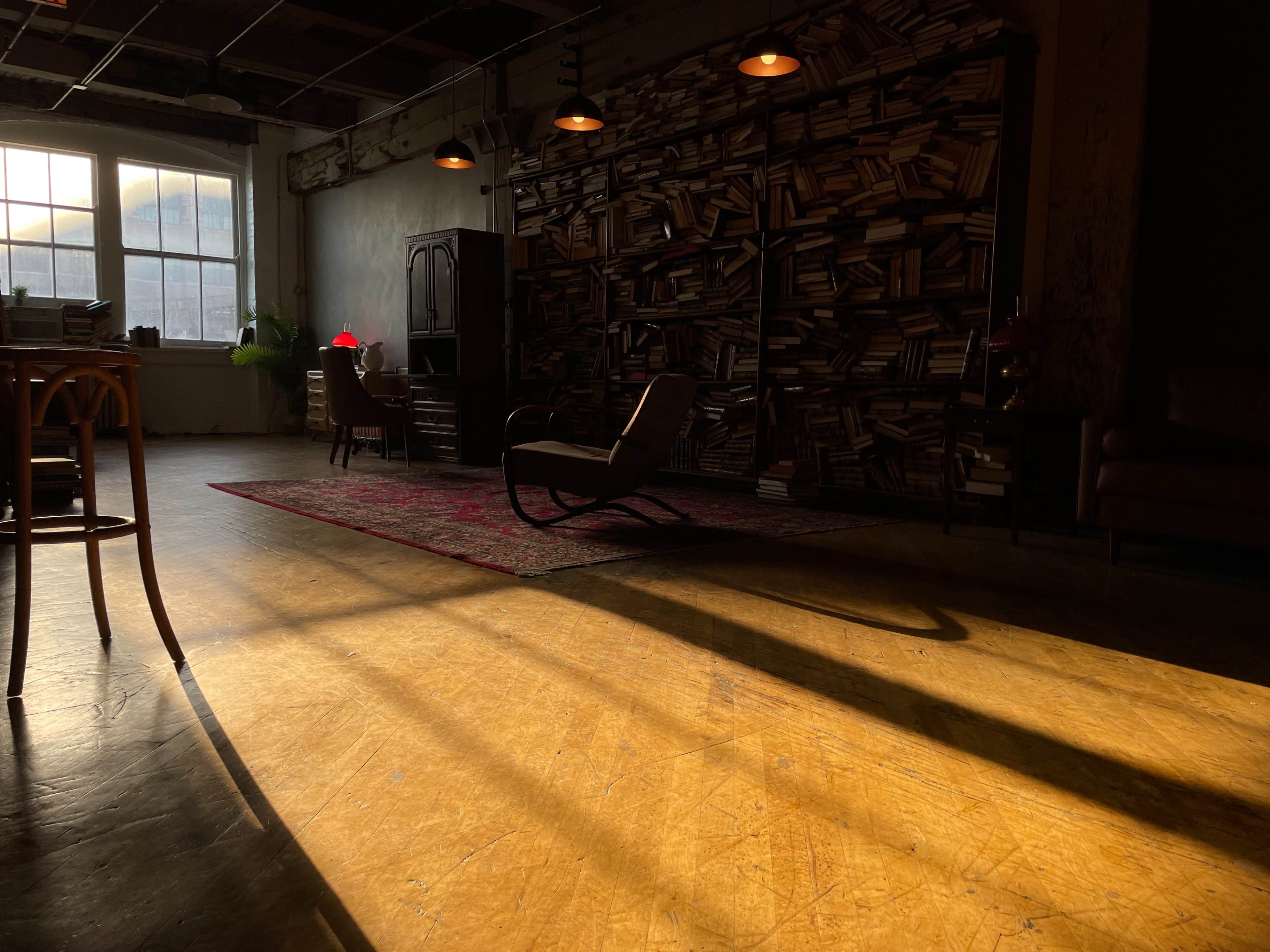 A cozy room with a wooden floor, a bookshelf filled with books on one wall, and a few pieces of furniture illuminated by sunlight streaming through a large window.