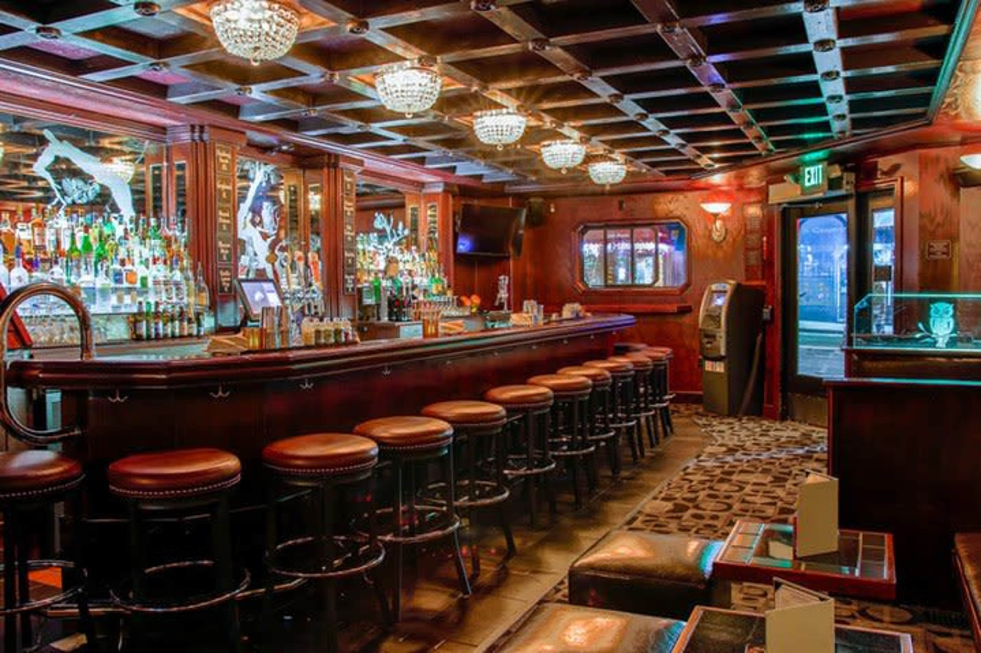 The image shows the interior of a bar with a long wooden counter, bar stools lined up in front, and shelves stocked with various bottles of liquor.