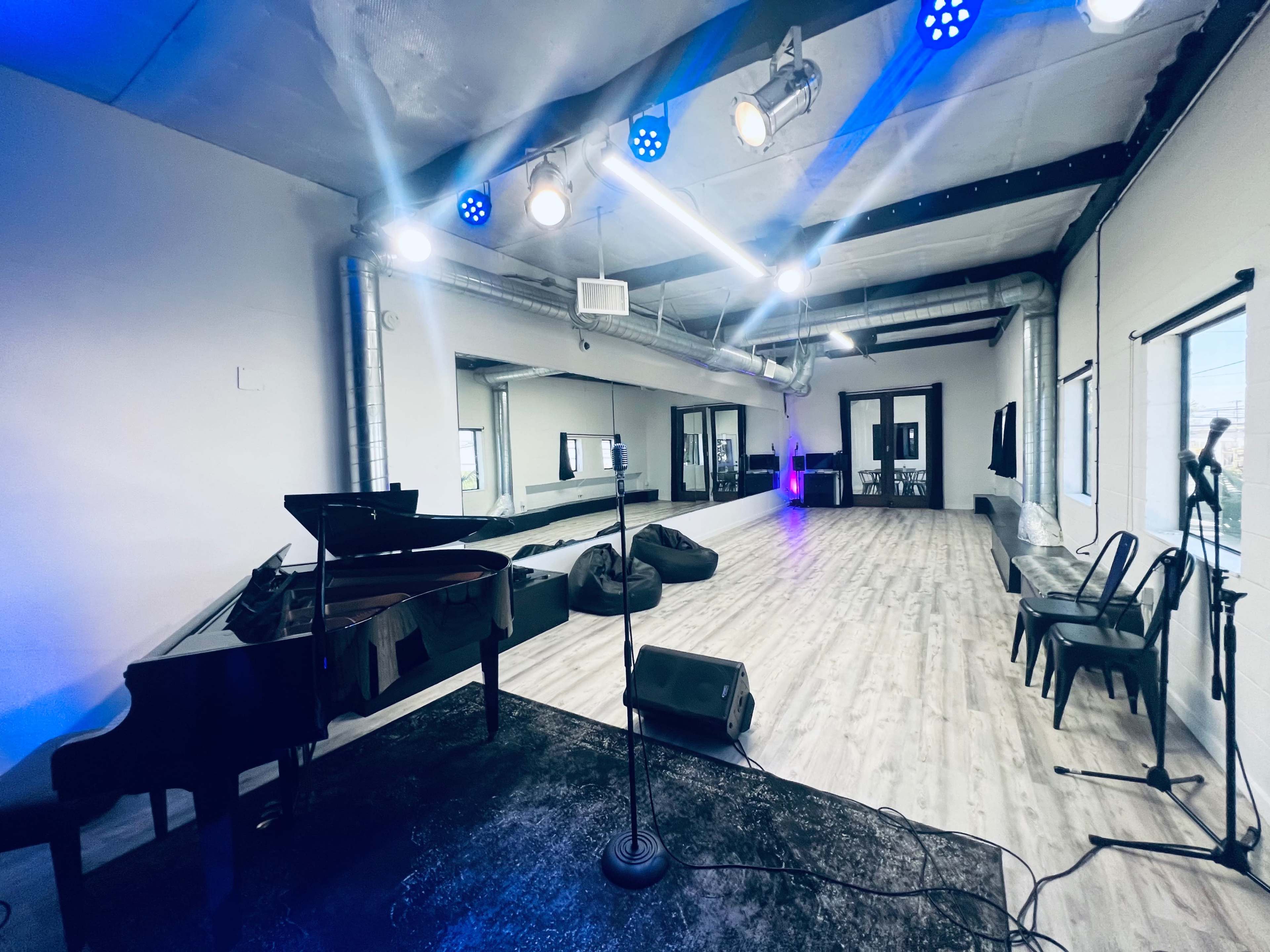 A spacious, well-lit performance room with a grand piano, a microphone stand, and several chairs arranged around a wooden floor.