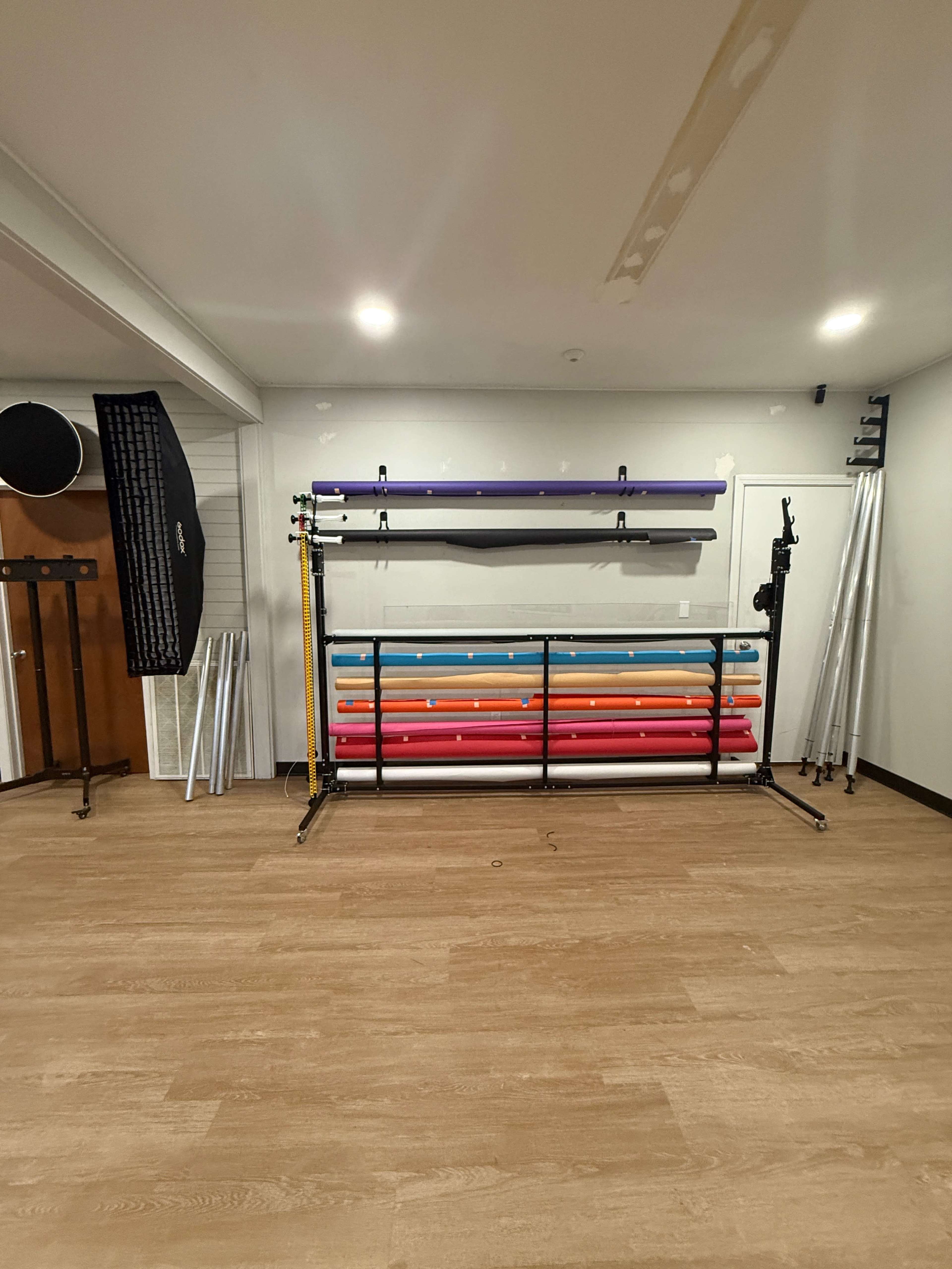 The image shows a room with wooden flooring, featuring a rack holding various colored poles and a backdrop support system on the wall.