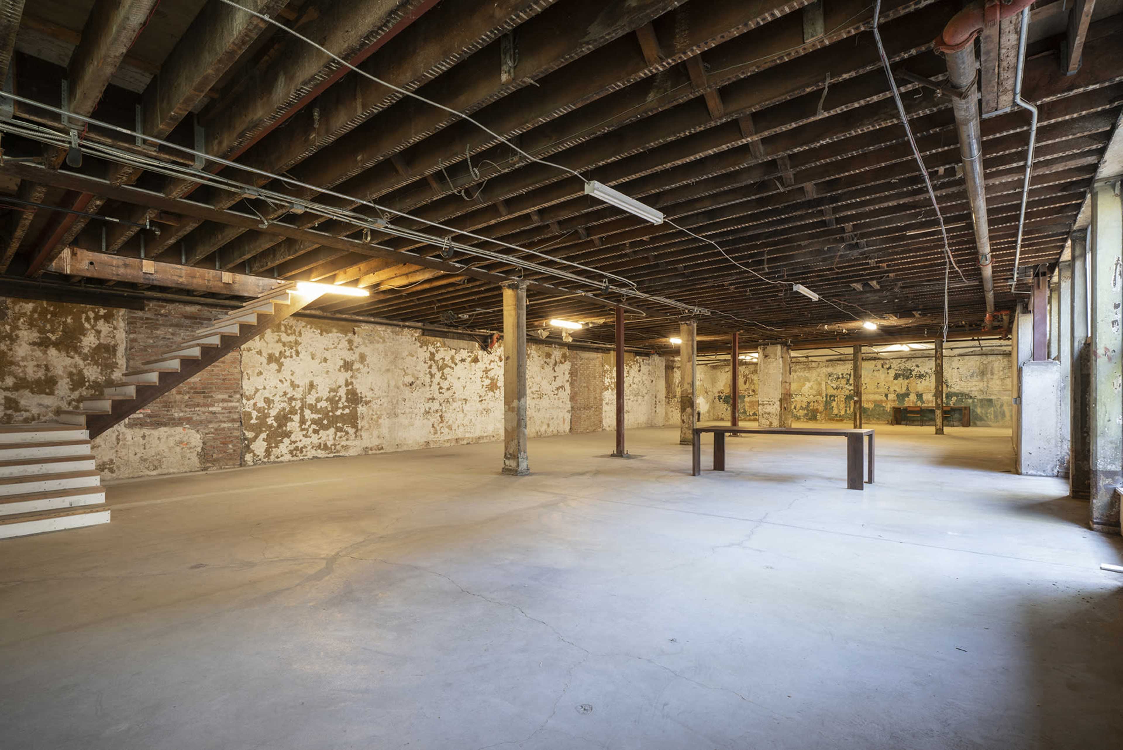 A spacious, empty basement with exposed beams, a concrete floor, and a wooden table in the center.