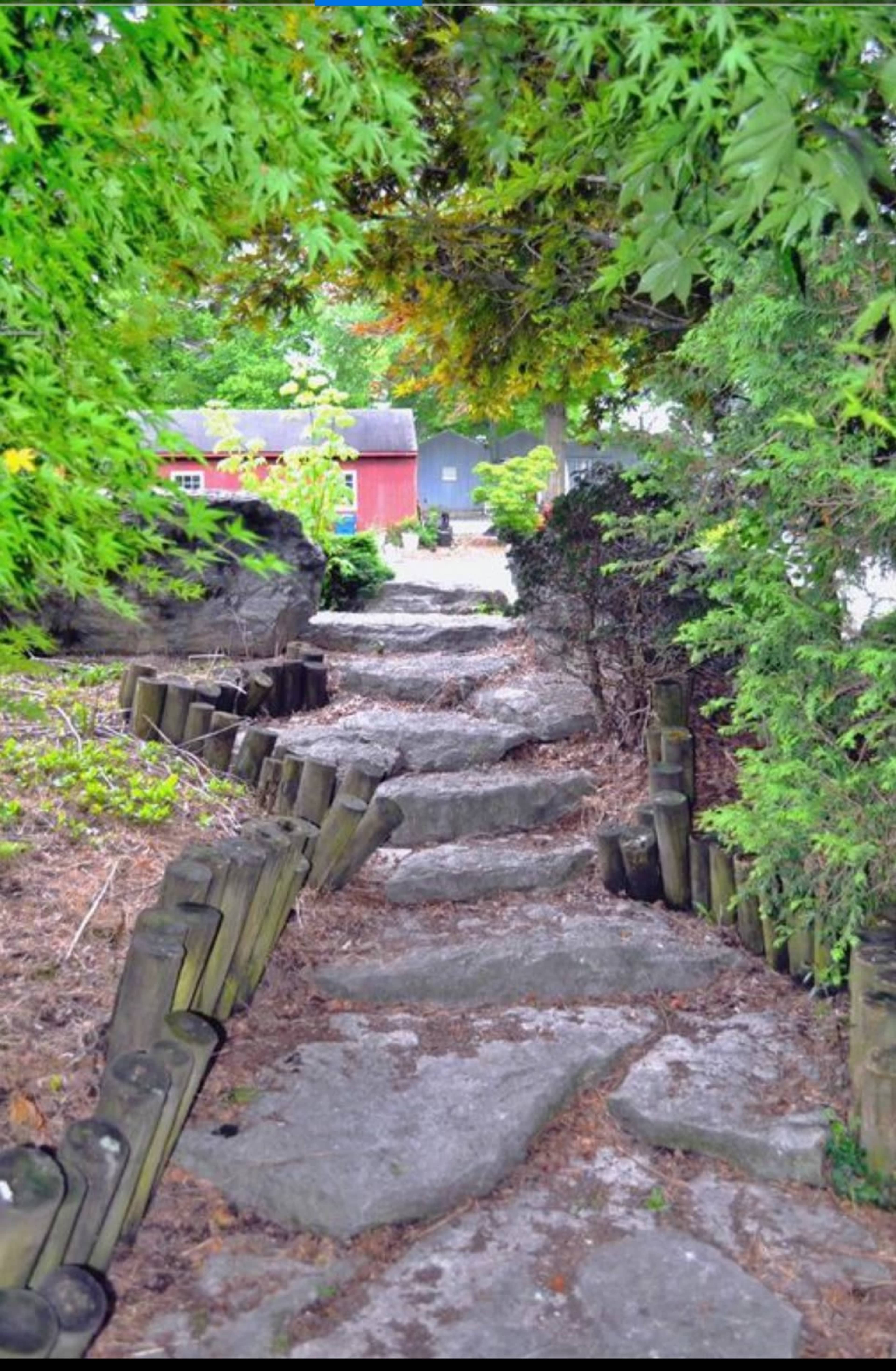 A stone pathway lined with wooden posts leads through a garden area with greenery and a glimpse of buildings in the background.