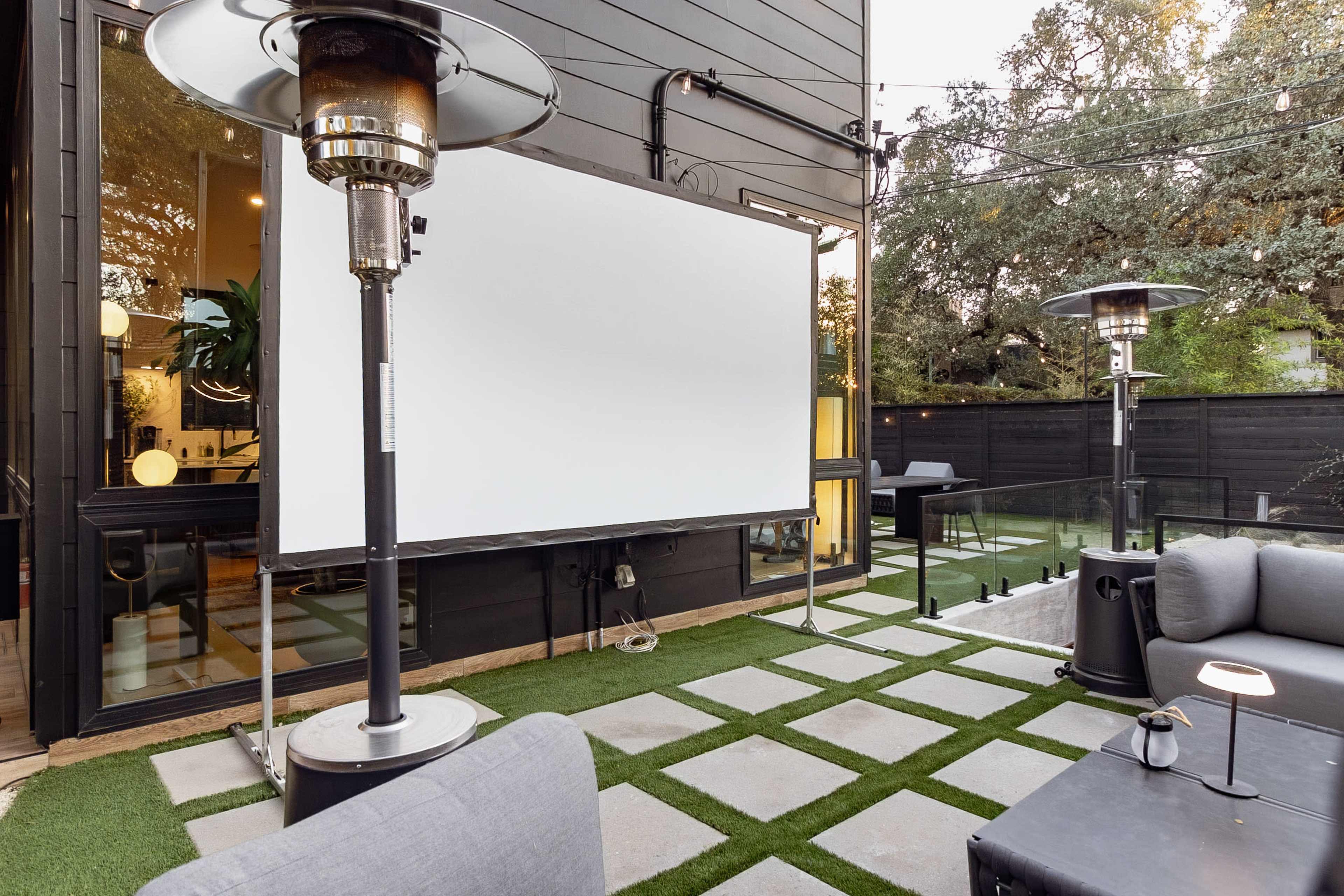 A backyard patio features a large blank projection screen surrounded by modern outdoor furniture and heating lamps on a grassy surface.