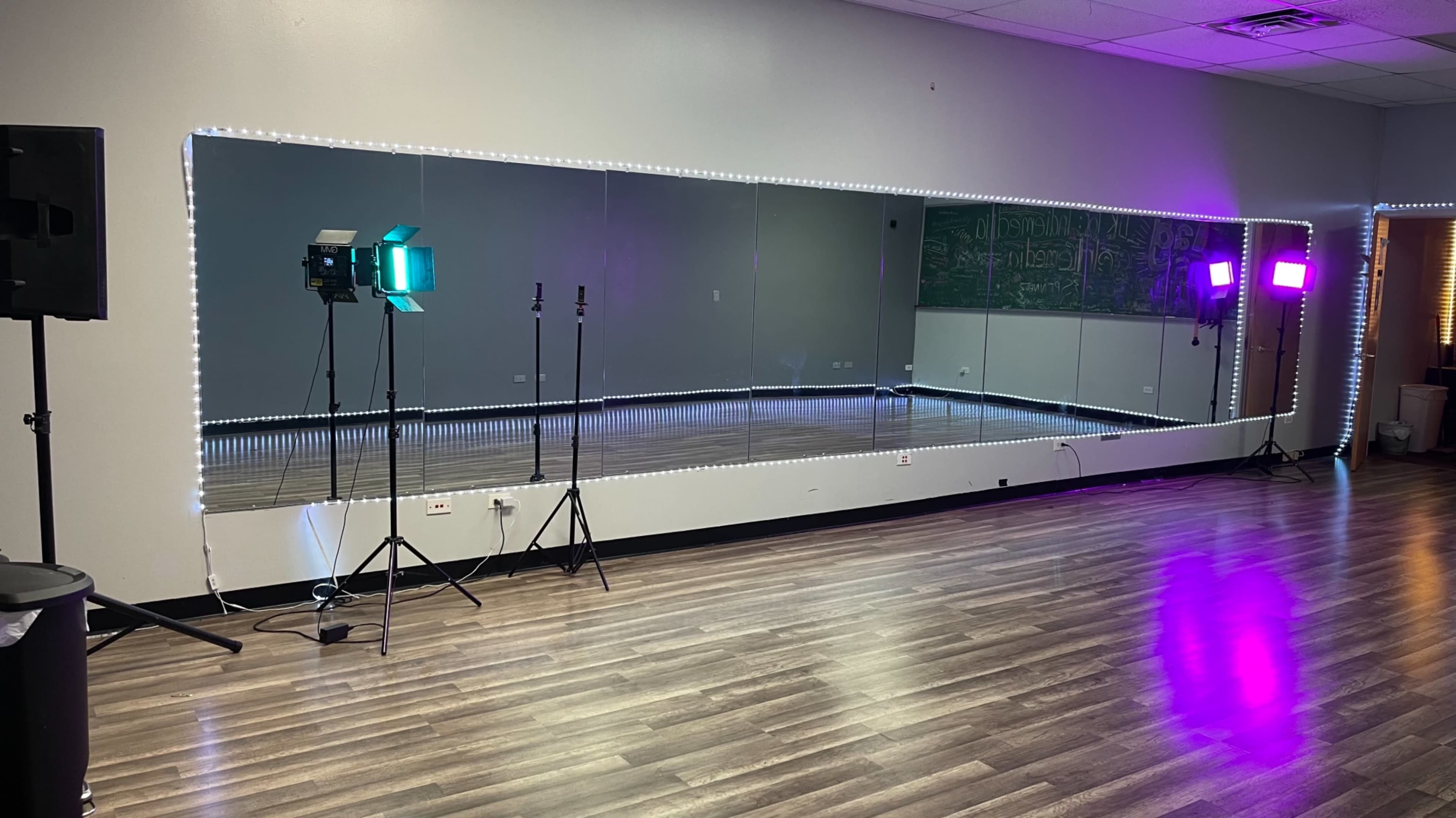 A spacious dance studio with a large mirror, lighting equipment on stands, and LED strip lights along the perimeter.