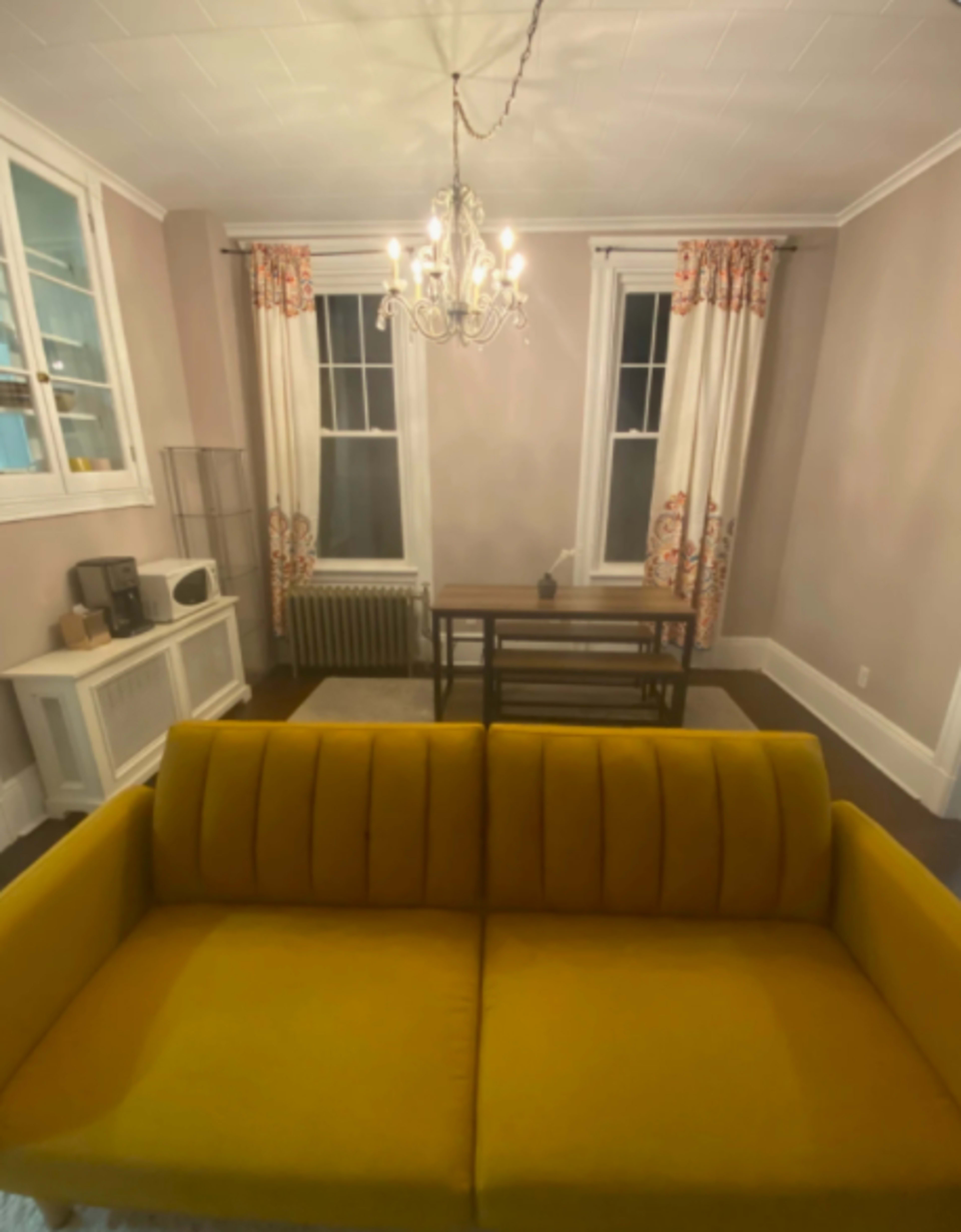 A yellow sofa faces a wooden table in a well-lit living room with light-colored walls and large windows adorned with curtains.