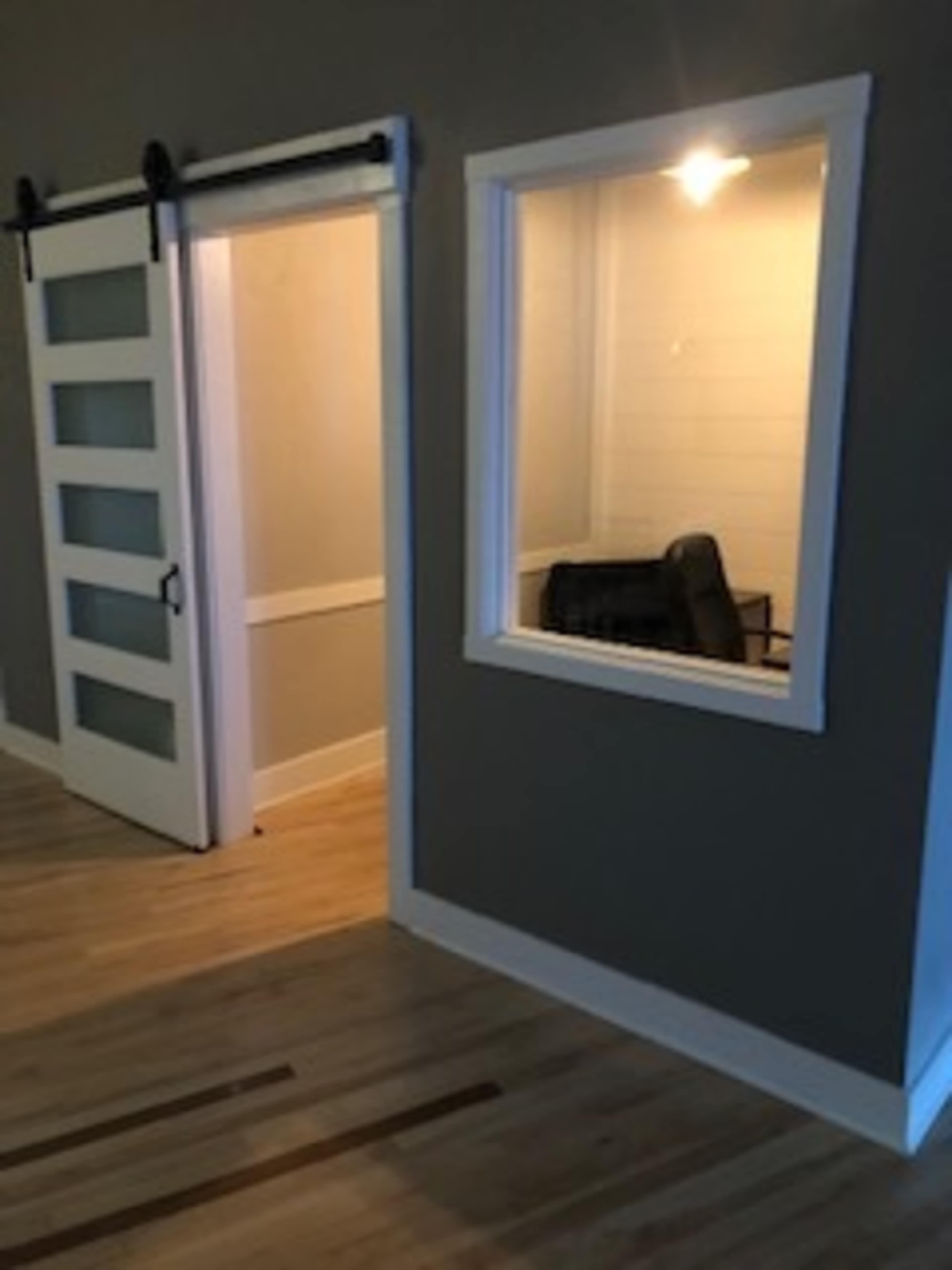 A room with a sliding barn door on the left and a window revealing a small, empty room with a black chair on the right.