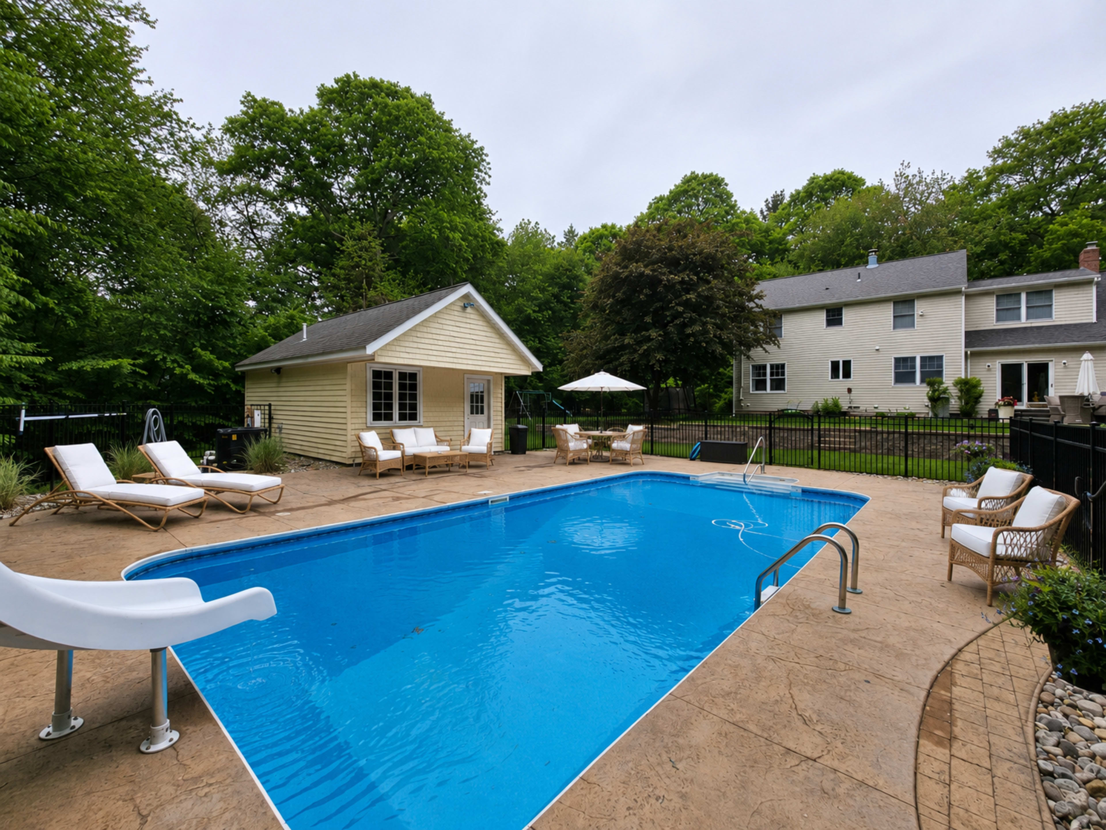 The image shows a rectangular swimming pool surrounded by stone paving, lounge chairs, and two houses in a green, wooded area.