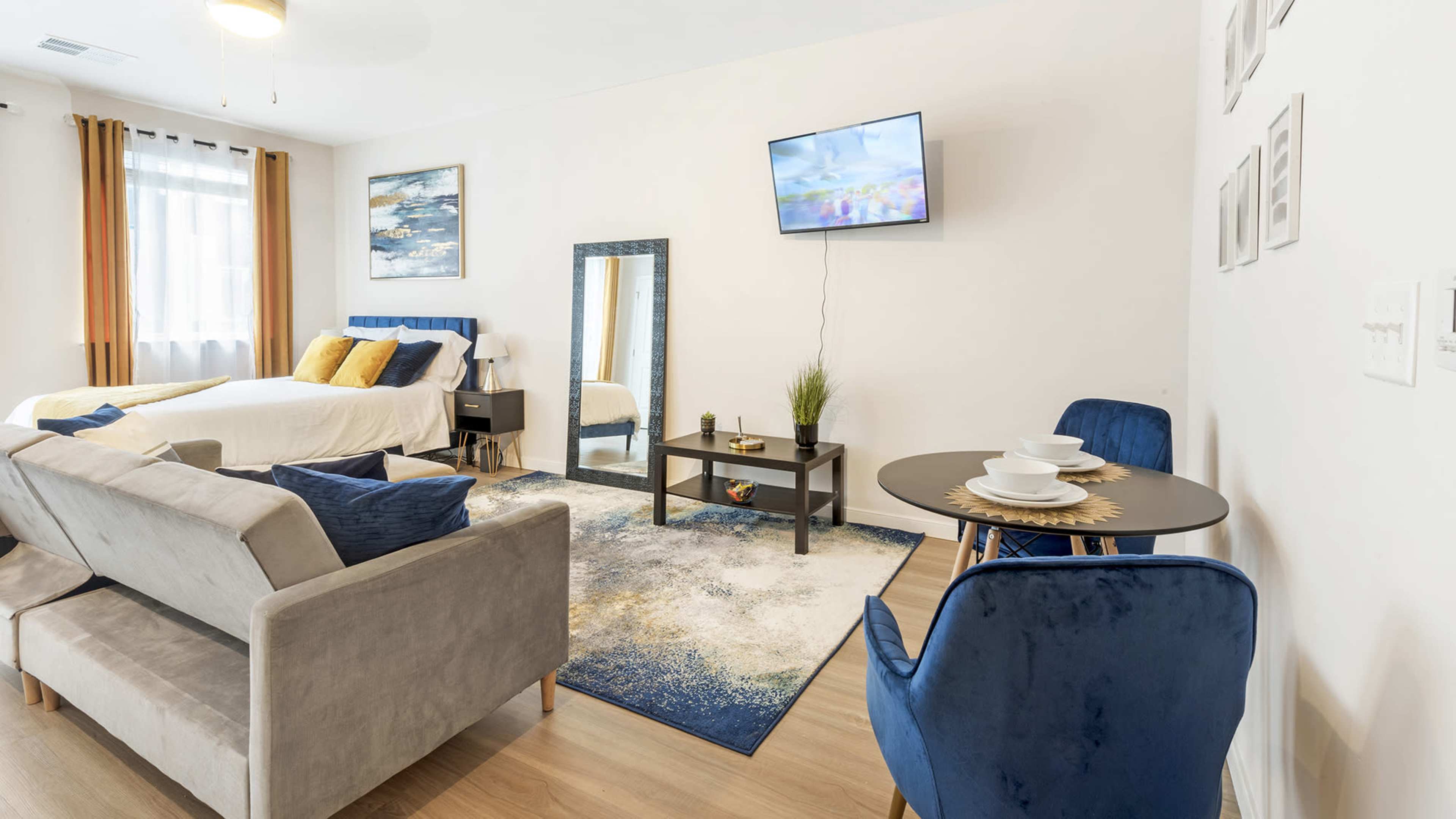 The image shows a modern studio apartment with a bed, a television on the wall, a seating area, and a dining table set for two.