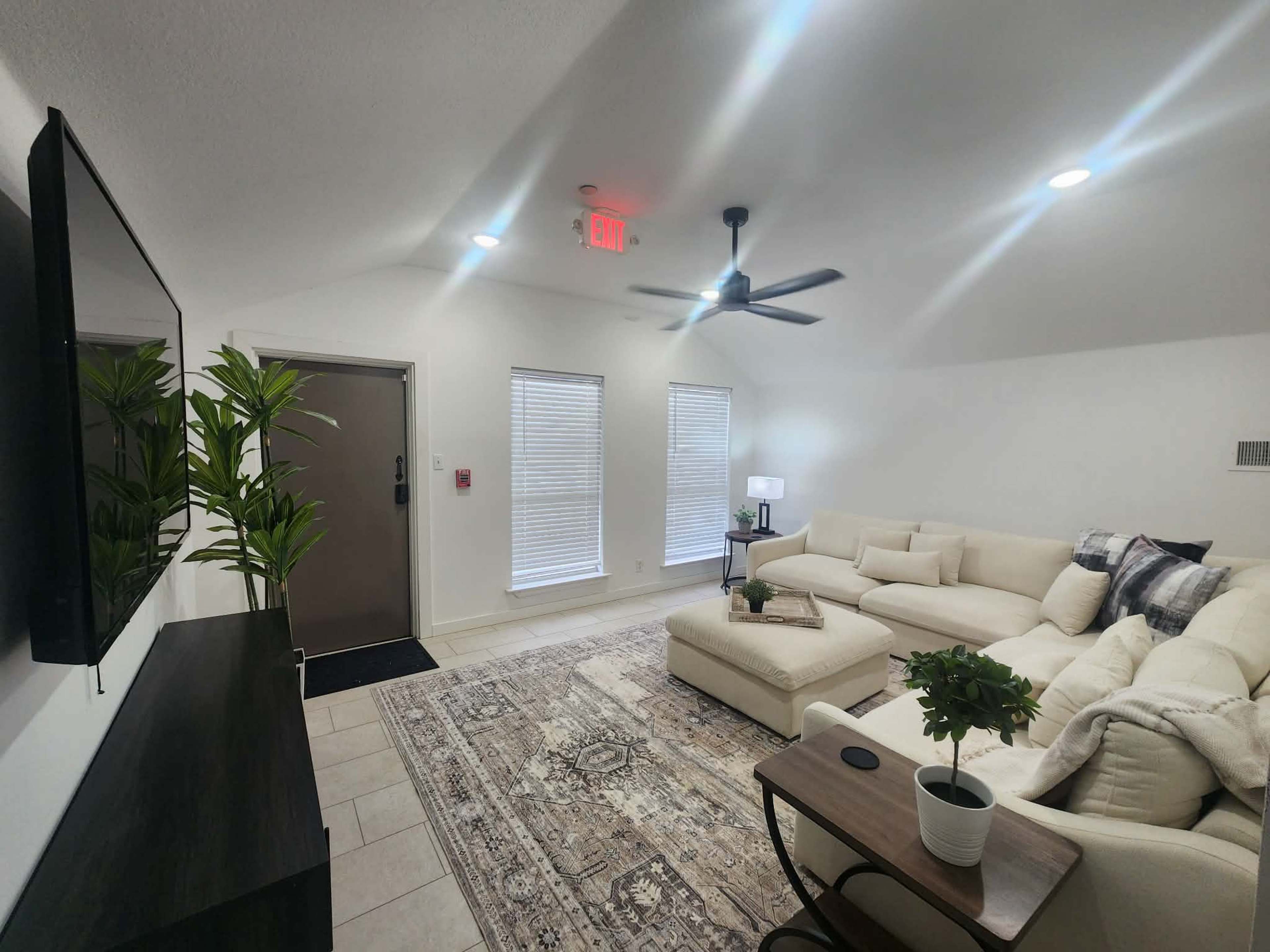 The image shows a modern living room with a beige sectional sofa, a coffee table, a television mounted on the wall, and a door leading outside, complemented by two windows and a ceiling fan.