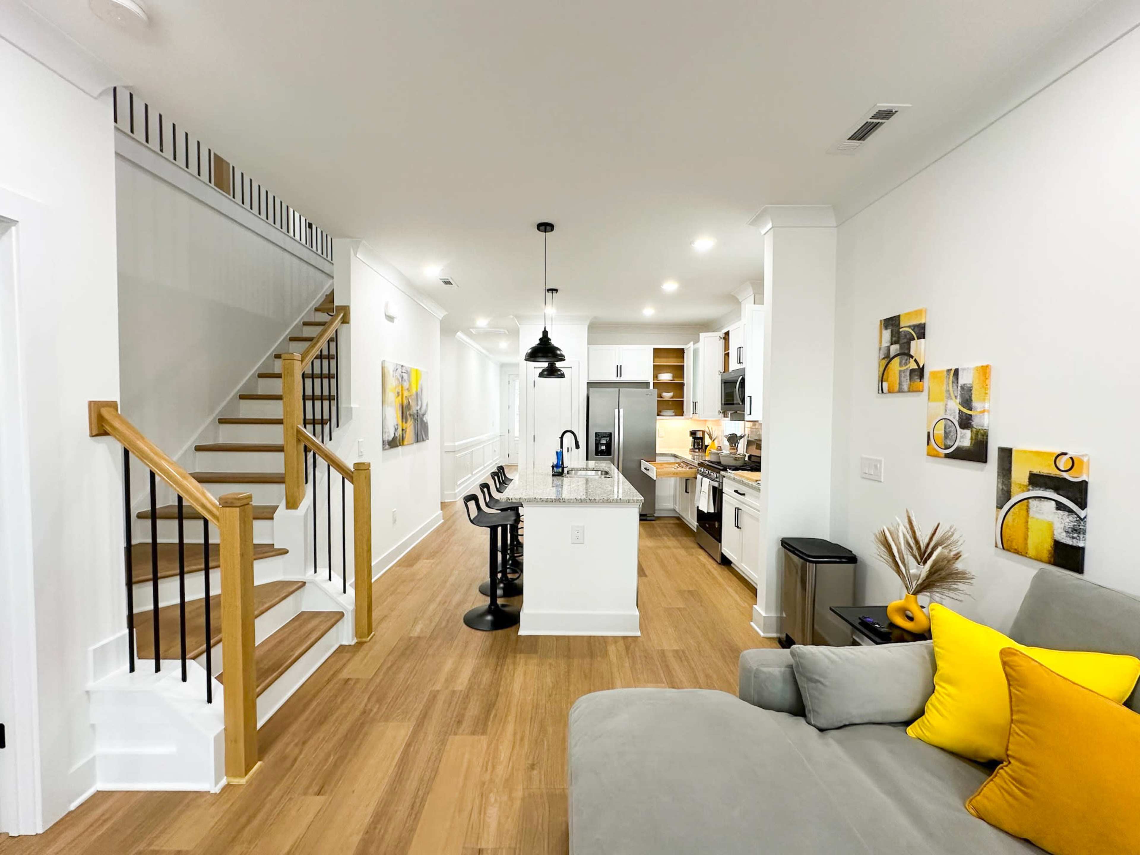A modern living space featuring a kitchen with an island, a staircase, and a sofa with yellow accents.