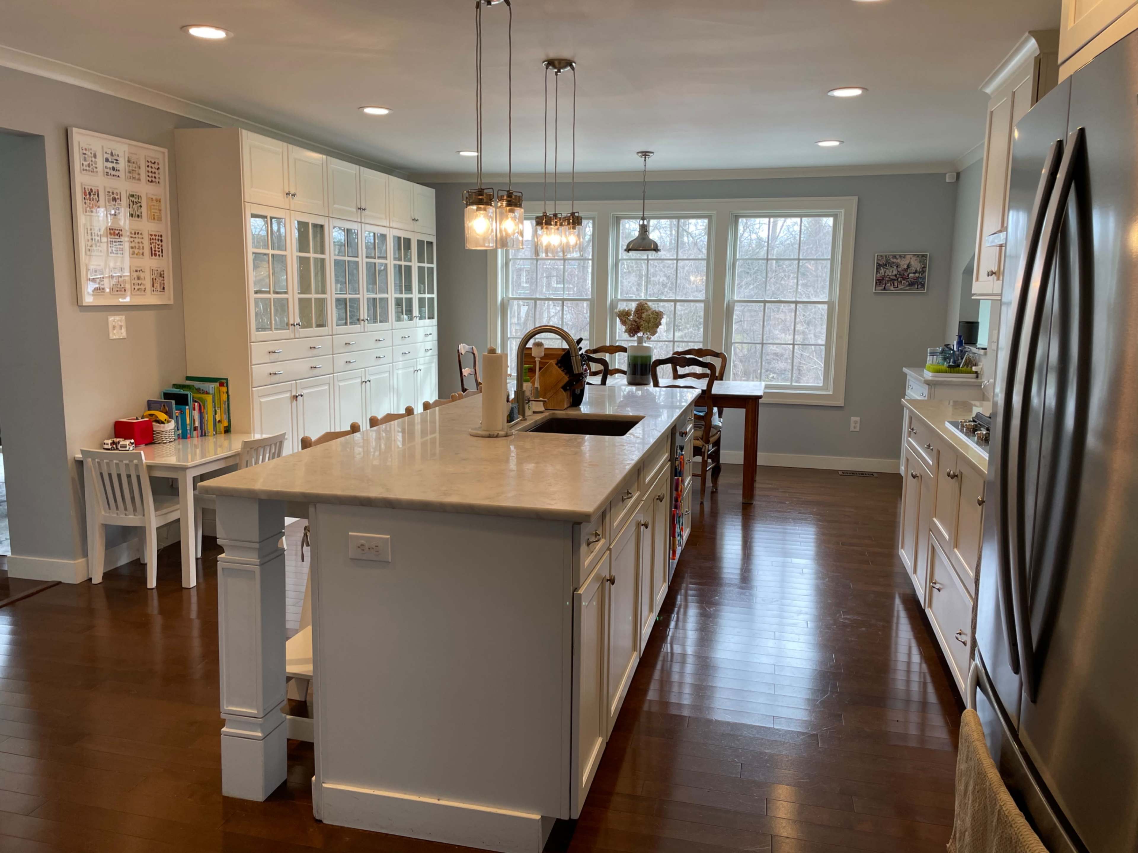 The image shows a modern kitchen with a central island, white cabinetry, and a dining area near large windows.