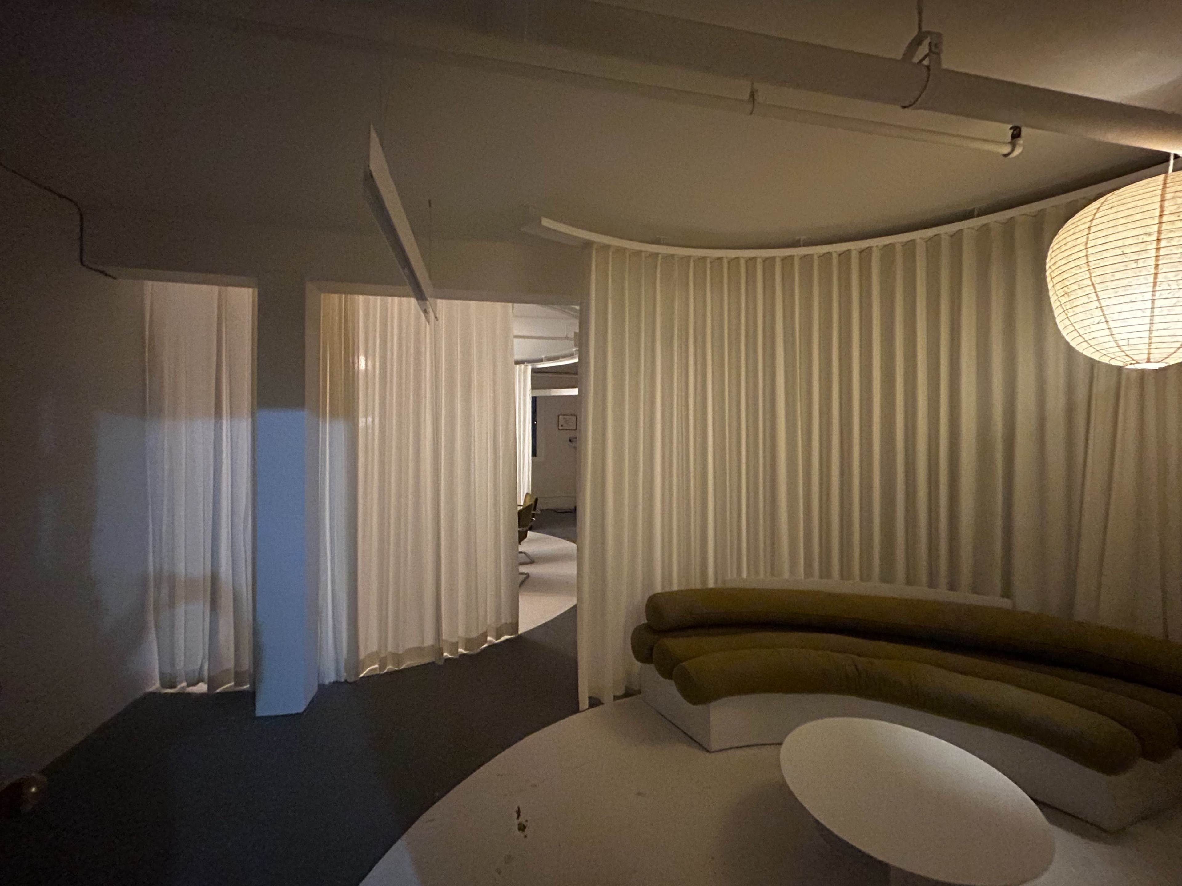 The image shows a softly lit modern interior space featuring a curved seating area, white drapery, and a round table under a large pendant light.