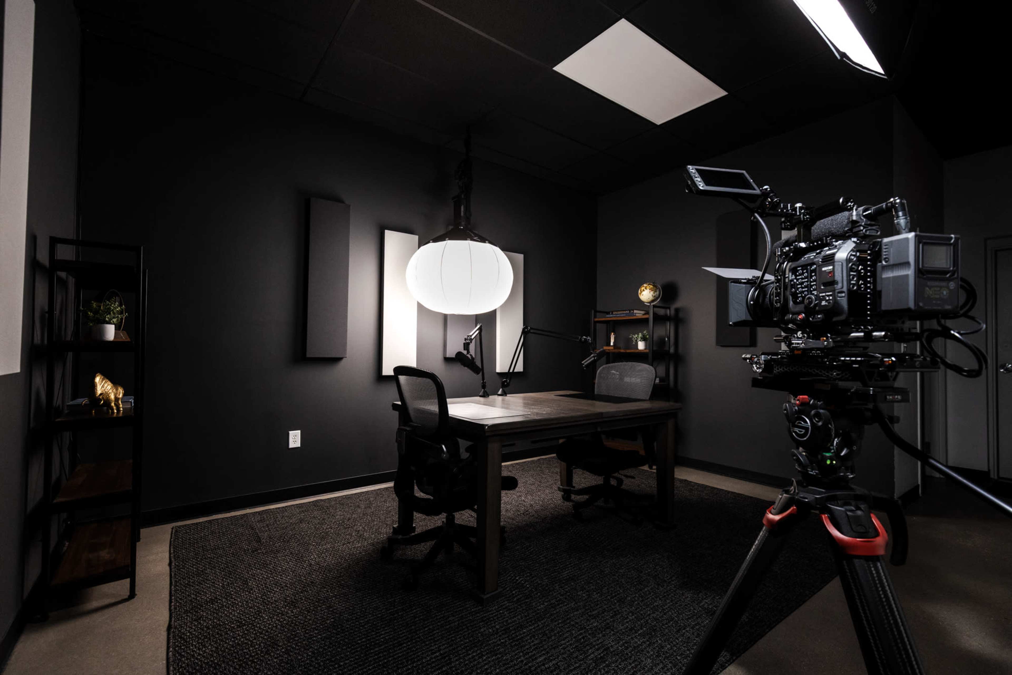 A video camera is positioned on a tripod facing a dark office space featuring a table, chairs, and overhead lighting.