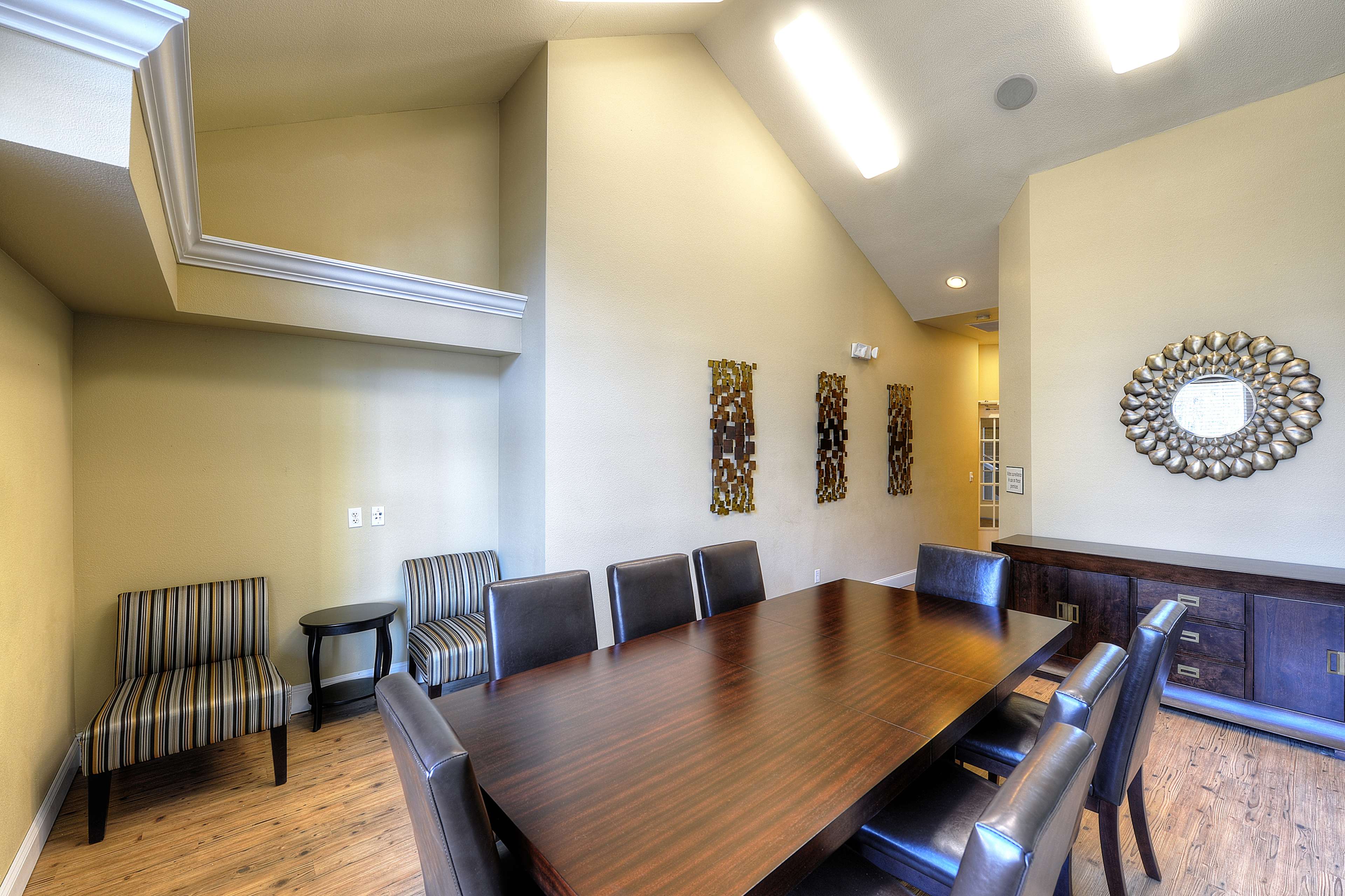 A meeting room is set up with a long dark wooden table surrounded by chairs, with additional seating and decorative wall art in a light-colored space.